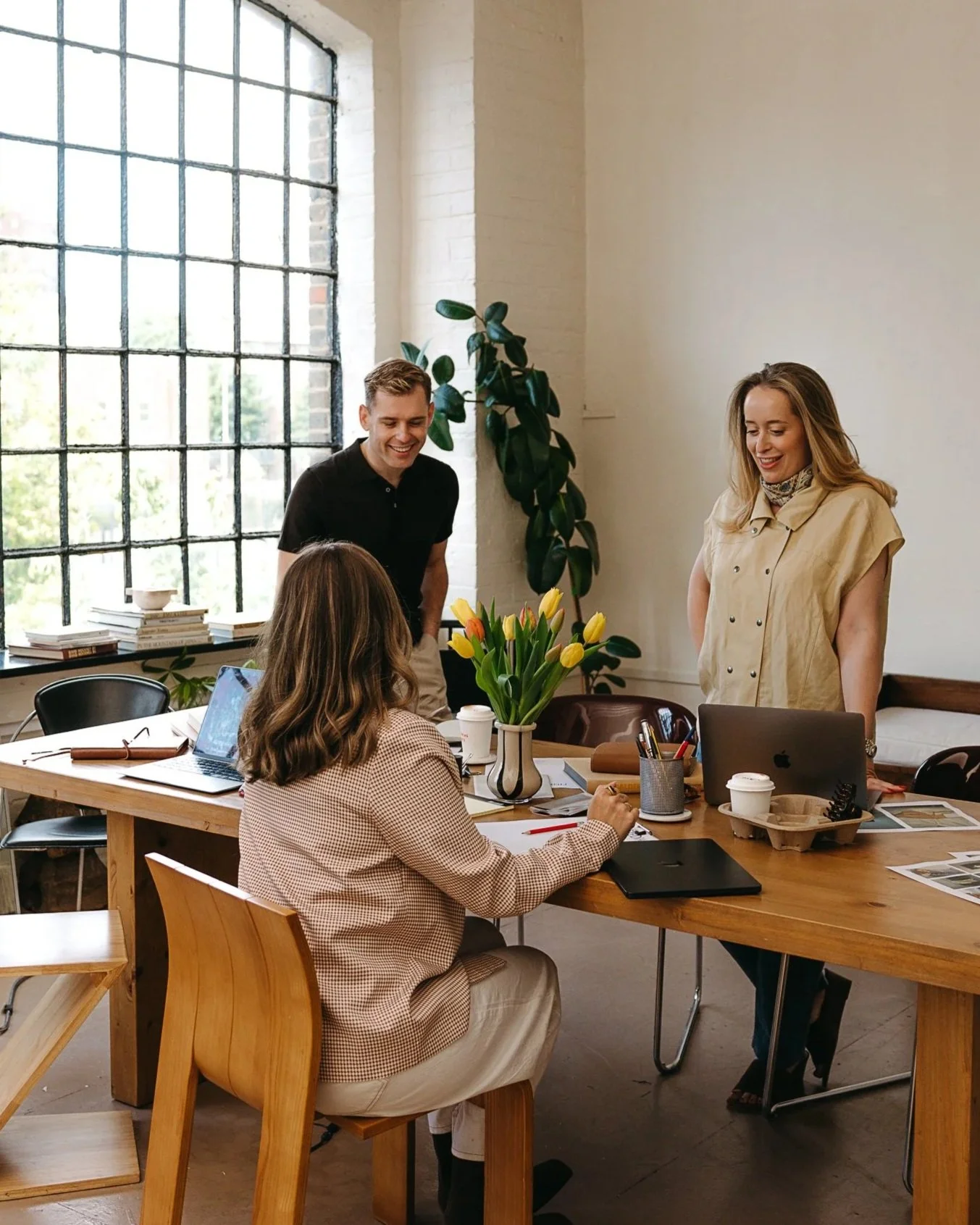 Finery Media team. A group of four young adults working and socialising in a bright, modern office with large windows. Two women are sitting at a wooden table with laptops, notebooks, coffee cups, and a center piece of yellow tulips.