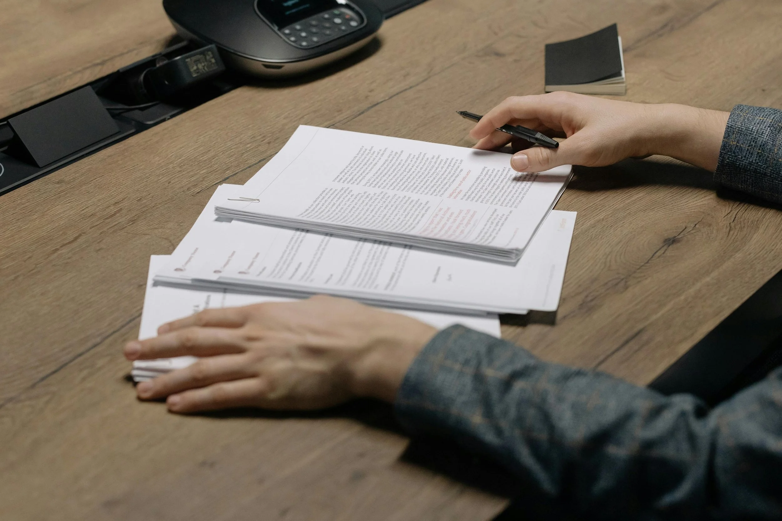 A person in a suit organizing and reviewing documents on a wooden desk, with a black office phone nearby.