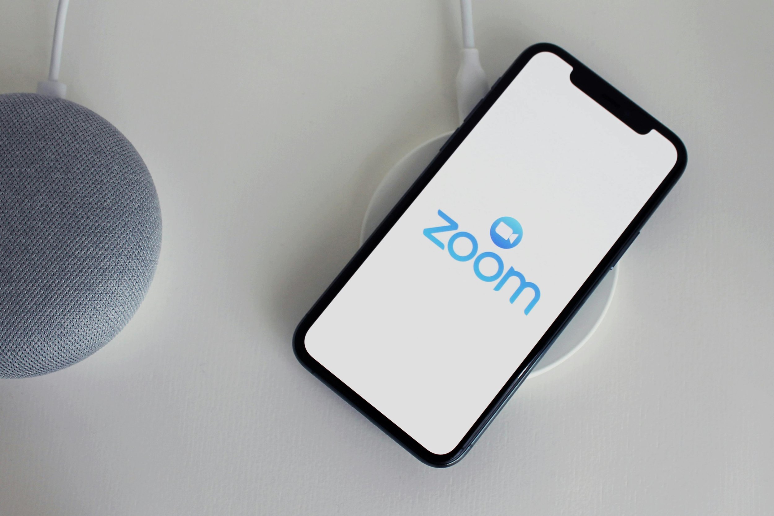 An iPhone displaying the Zoom logo on a charger, placed on a white surface next to a gray smart speaker.