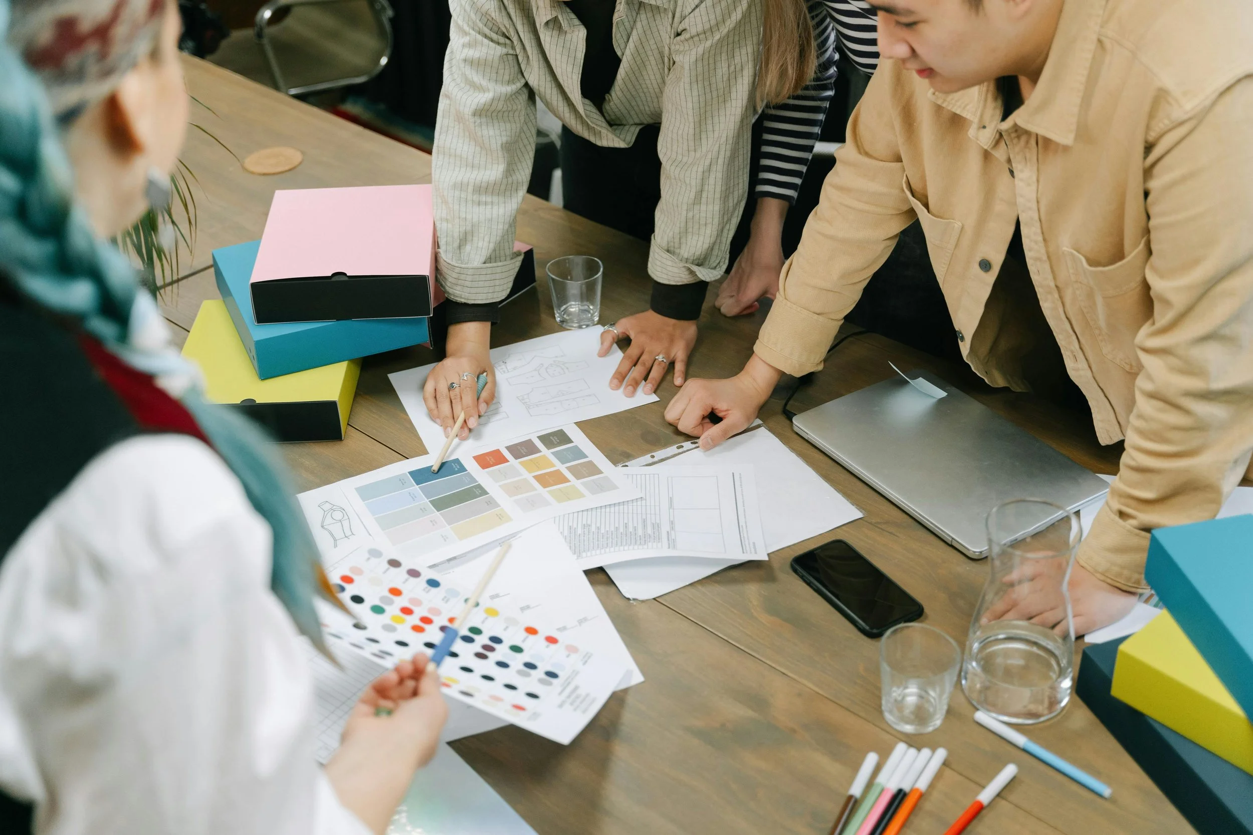 Team of people collaborating around a wooden table with color palettes, sketches, and documents, discussing project design.