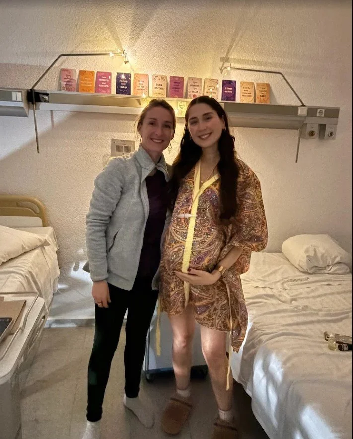 Two women standing in a hospital room, smiling, with hospital beds on either side, and a shelf with colorful boxes or books above them.