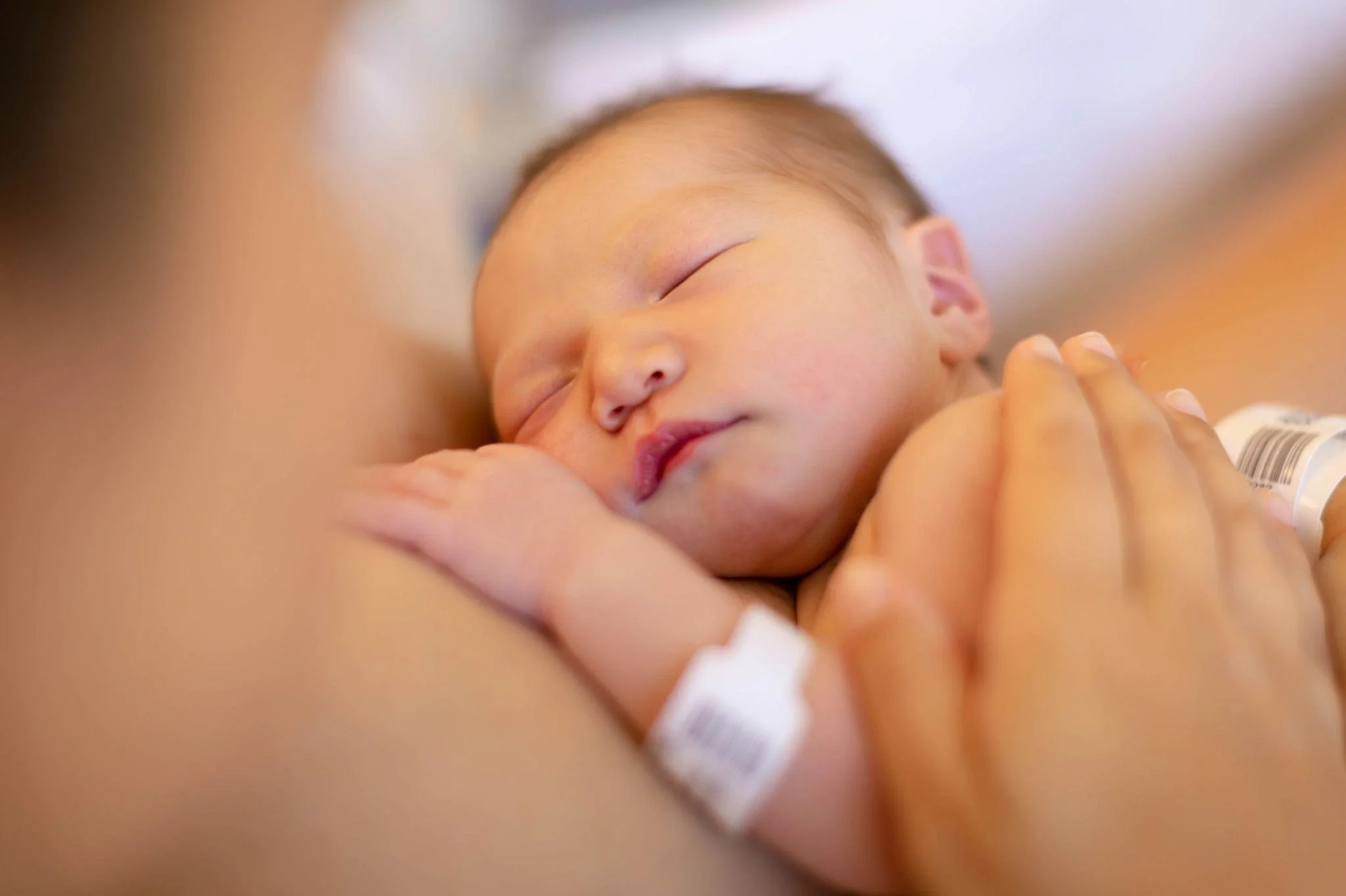 Close-up of a baby sleeping peacefully, being held gently by an adult, with a hospital wristband visible on the adult's wrist.