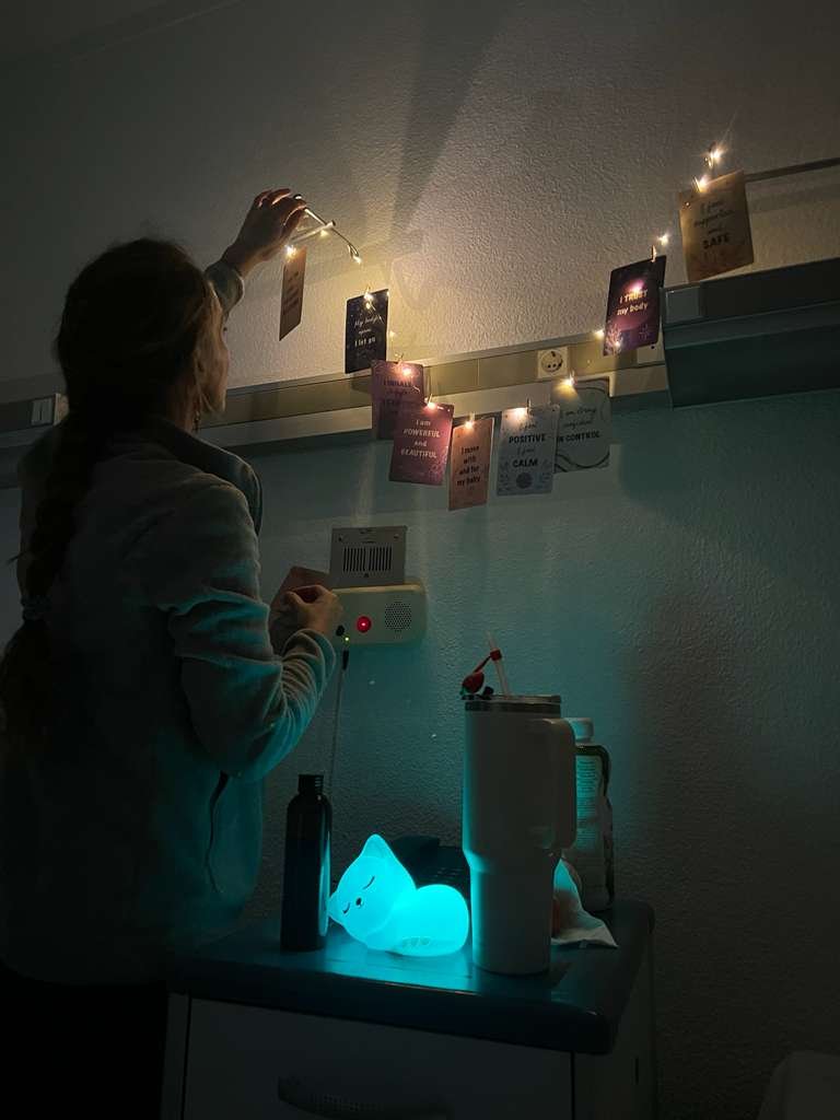 A woman hanging string lights above a wall with inspirational quotes and cards, with a glowing whale lamp on a table below.