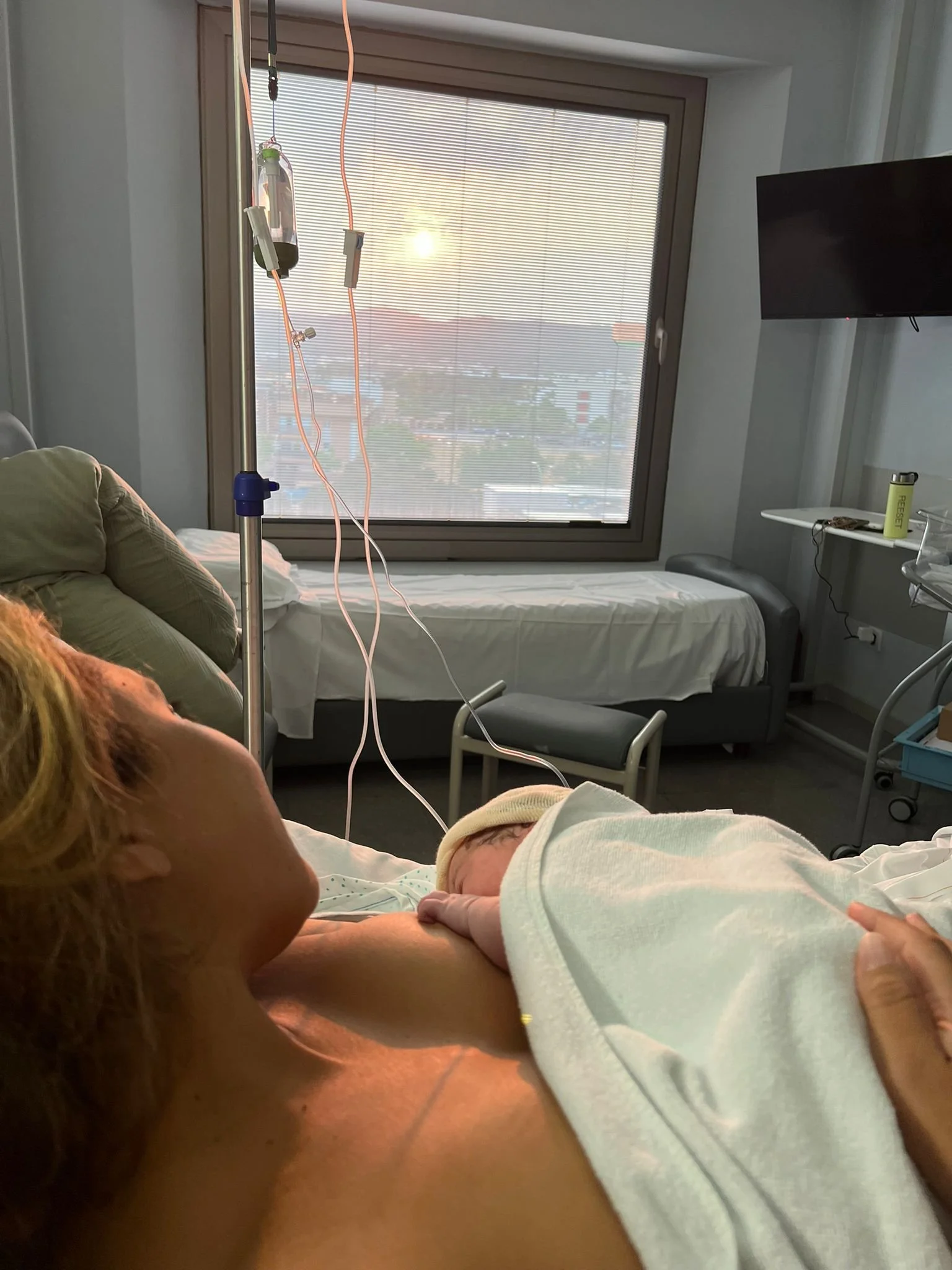 A woman lying on a hospital bed holding a newborn baby, with medical equipment and a window in the background.