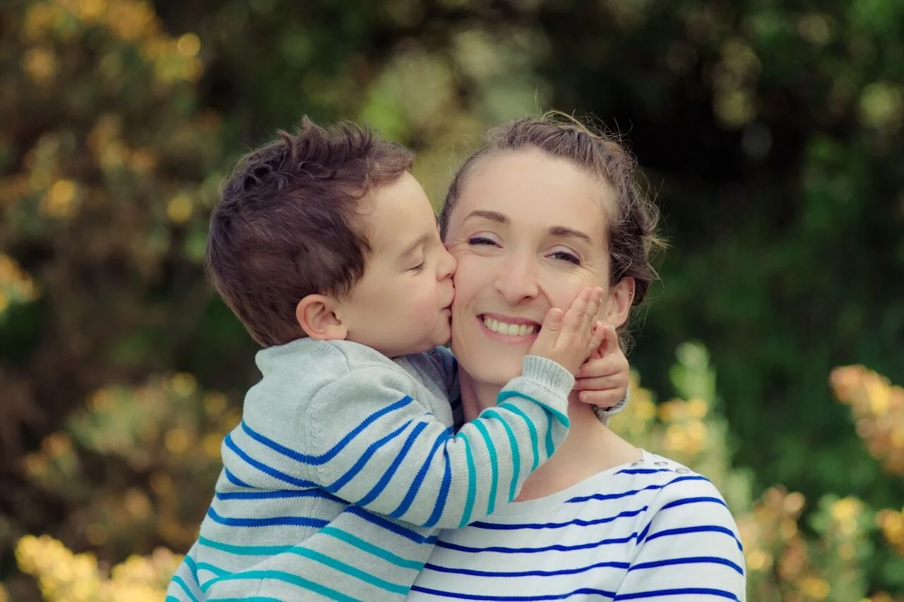 A woman with brown hair in a ponytail smiling with a young boy kissing her on the cheek outdoors with blurred green and yellow trees in the background