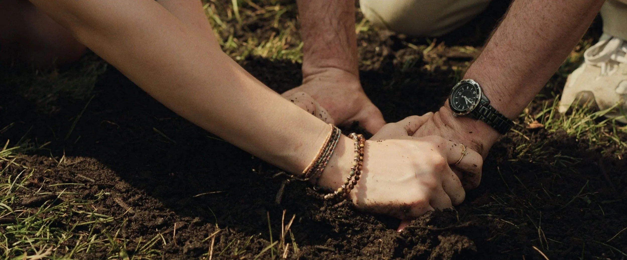 Two people are planting a seed in the soil, holding hands and wearing casual clothes and watches, with one person wearing bracelets.