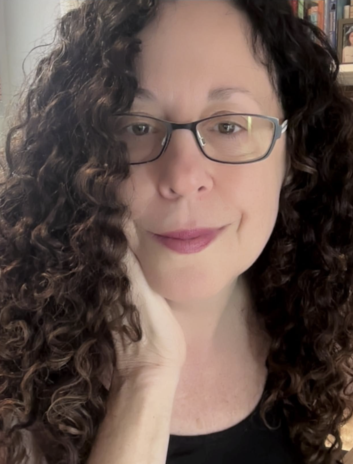 A woman with curly brown hair, glasses, and light makeup, wearing a black top and a beige scarf, smiling gently at the camera in a cozy indoor setting with bookshelves in the background.