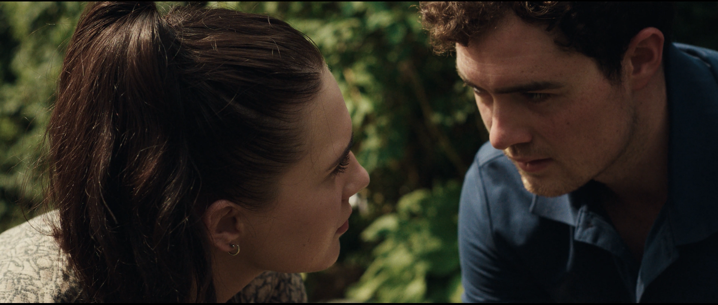 A close-up of a woman and a man facing each other outdoors, with the woman slightly looking up at the man. The background features green foliage.