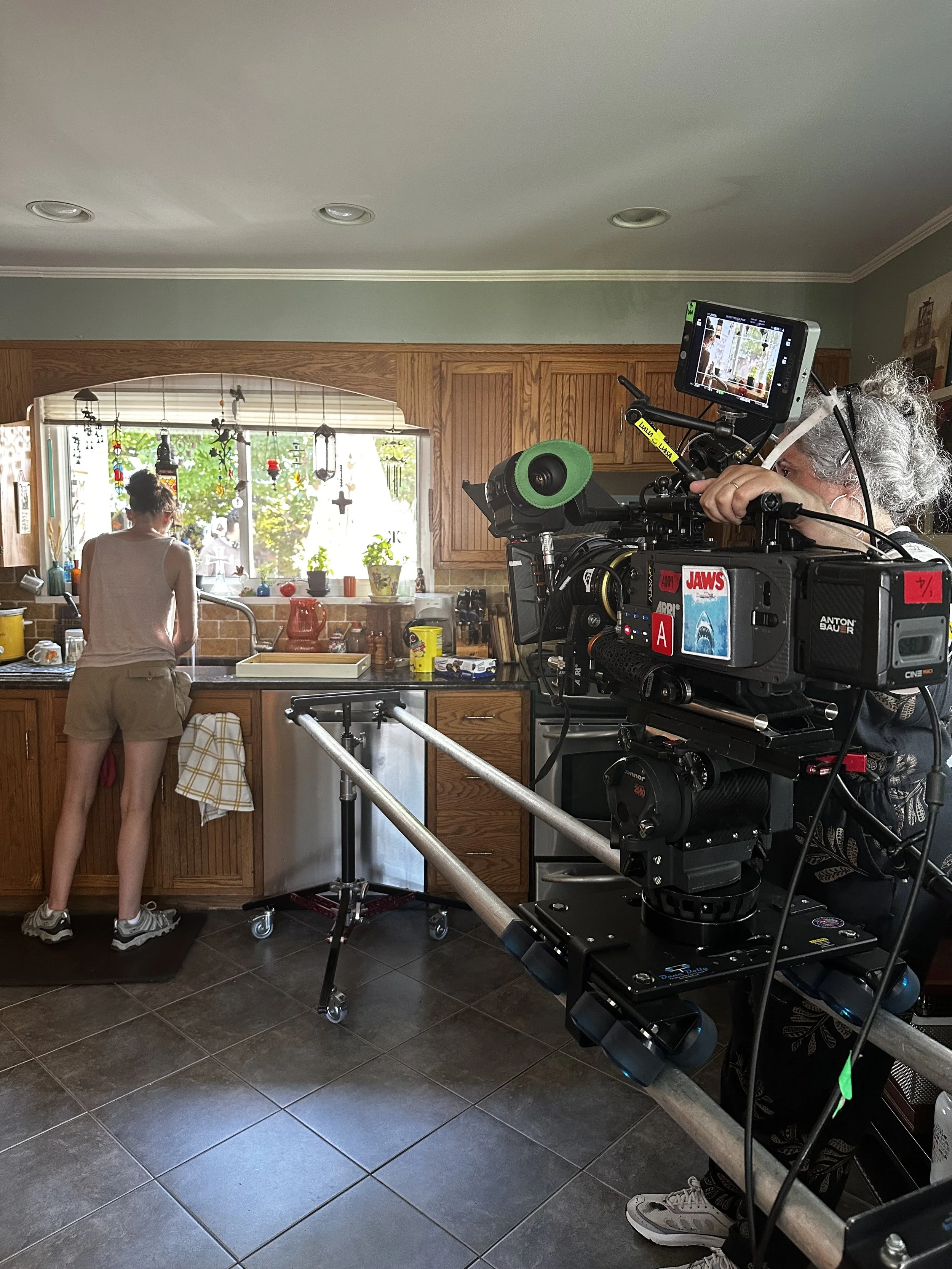 Behind the scenes of a kitchen scene filming, a person stands at the sink while a camera crew films with a professional camera setup near the counter.