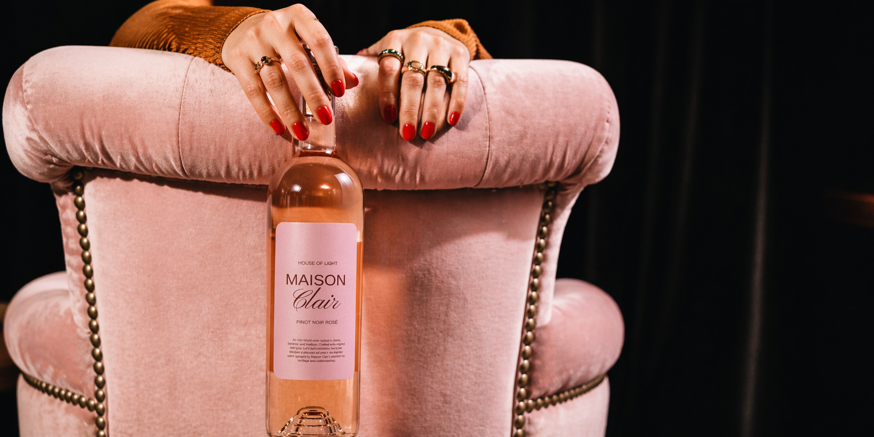 A person's hands with red nails and multiple rings rest on a pink velvet armchair, with a bottle of Maison Clair rosé wine placed in front of the chair.