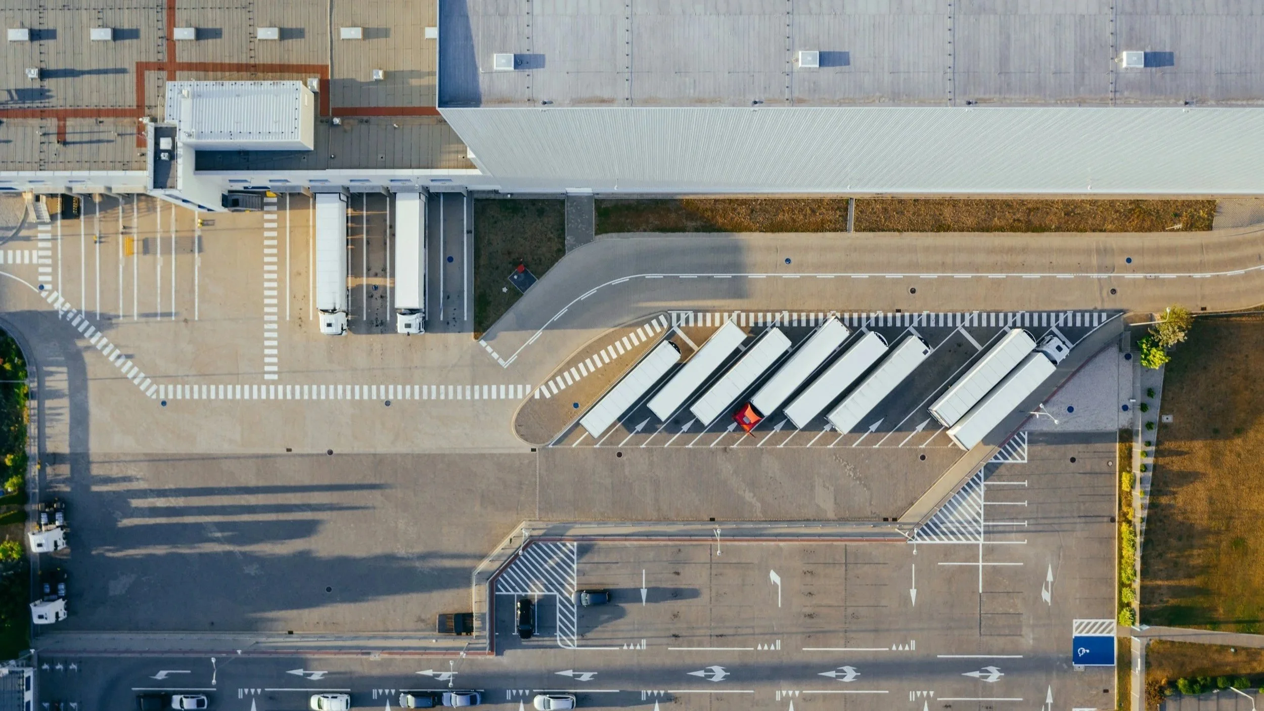 An aerial view of a modern building with a large, flat roof. There are multiple trucks parked in designated areas and a road with marked lanes and crosswalks surrounding the building.