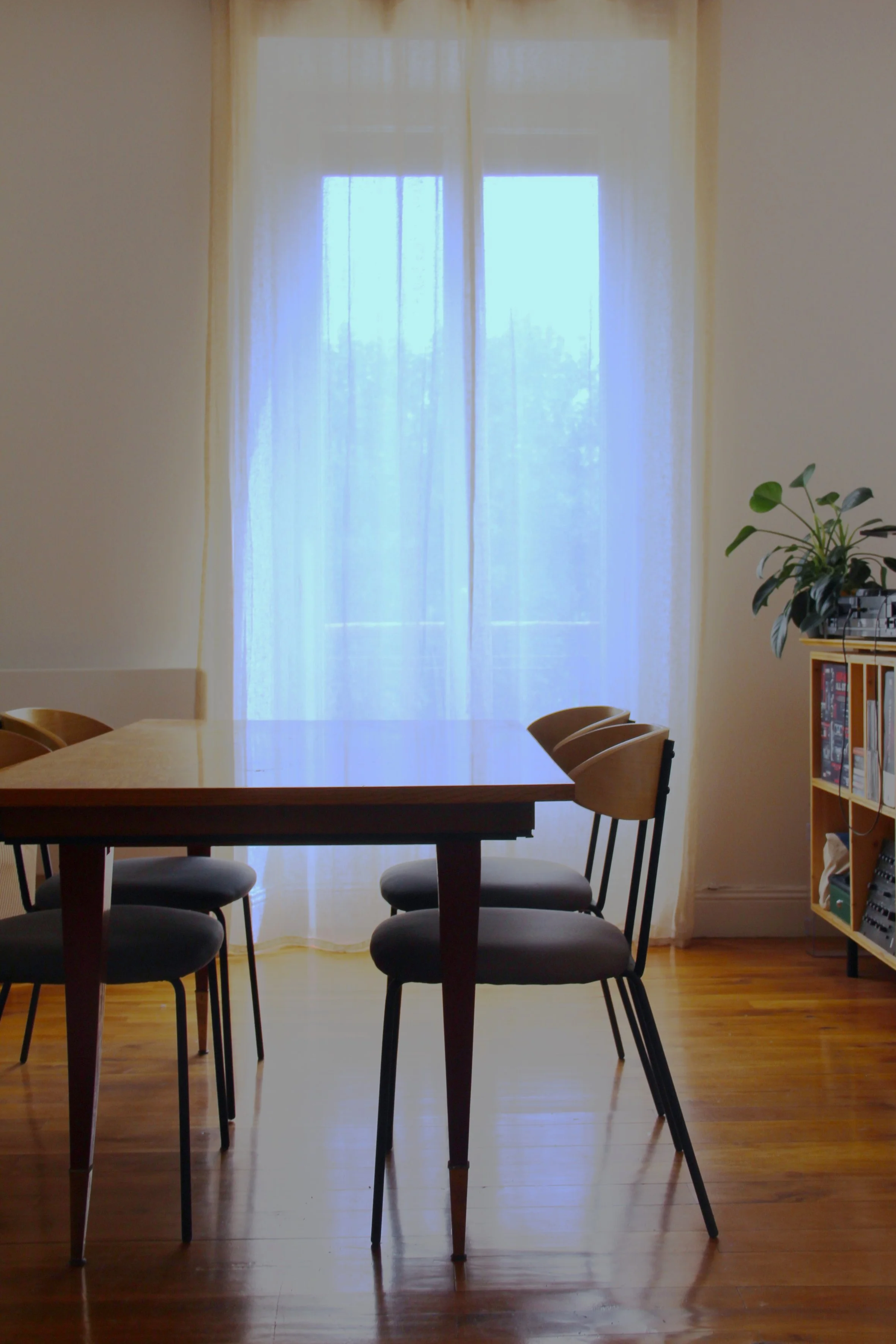 Une salle à manger avec une grande table en bois entourée de six chaises, une grande fenêtre avec des rideaux légers laissant passer la lumière et un meuble avec des livres et une plante verte.