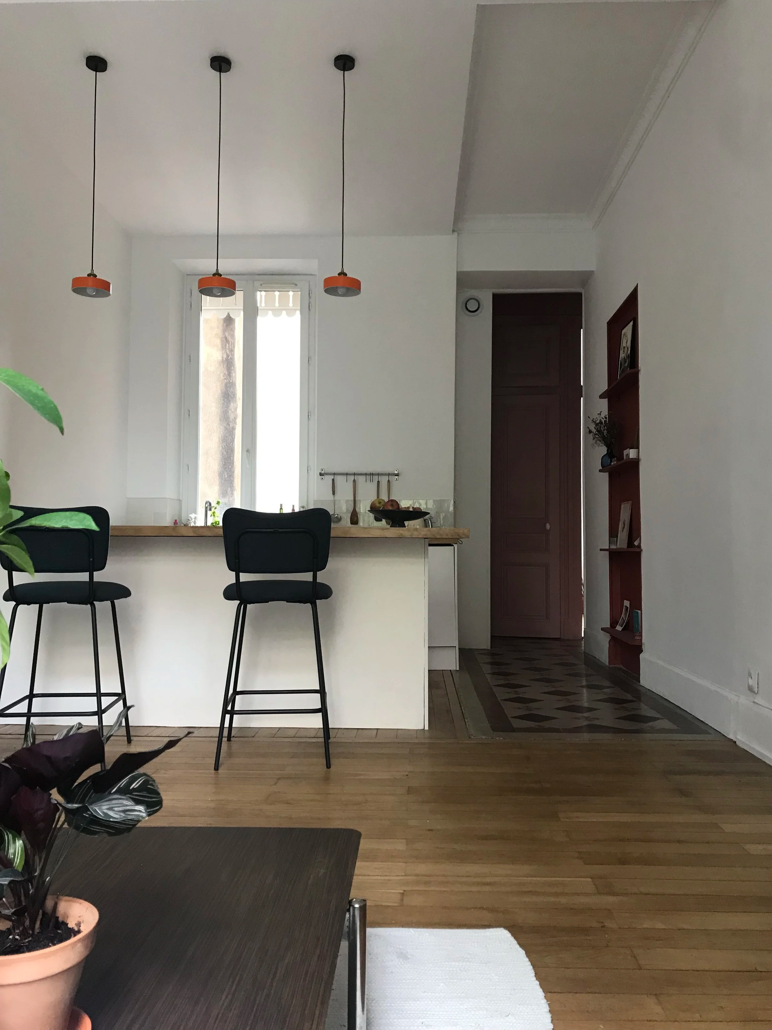 Une salle à manger avec un îlot central, une fenêtre laissant entrer de la lumière naturelle, une niche en bois rouge avec des éléments de décoration, un sol en parquet, un plafond blanc avec trois suspensions oranges.