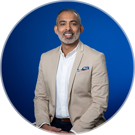 A middle-aged man with a gray beard, wearing a beige blazer over a white shirt, smiling in front of a blue background.