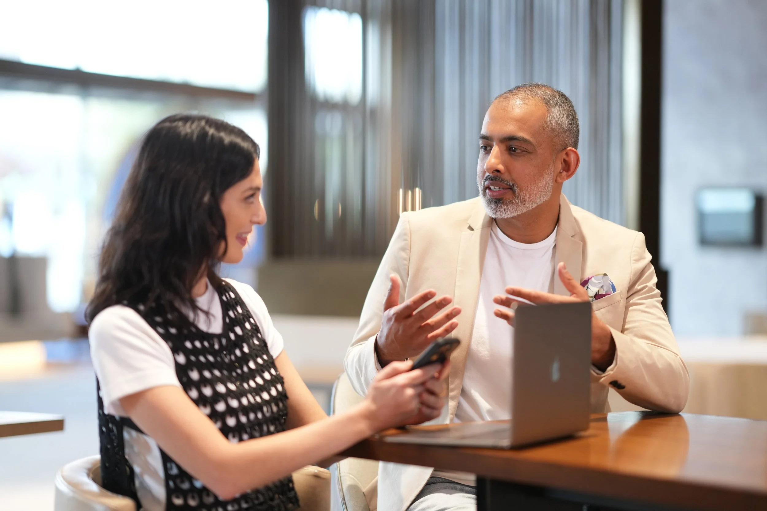 A man and woman engaged in a conversation at a cafe or restaurant, sitting at a wooden table with a modern, softly lit interior in the background.