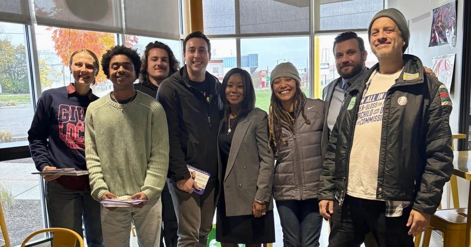 Group of nine diverse activists standing indoors, smiling for the photo, with large windows and an outdoor parking lot visible behind them.