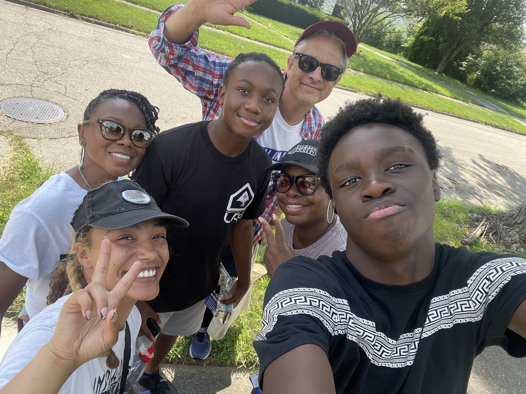 Group of volunteers for Mayor Shenise Turner-Sloss taking a selfie outdoors on a sunny day, standing on a sidewalk near green grass and trees.
