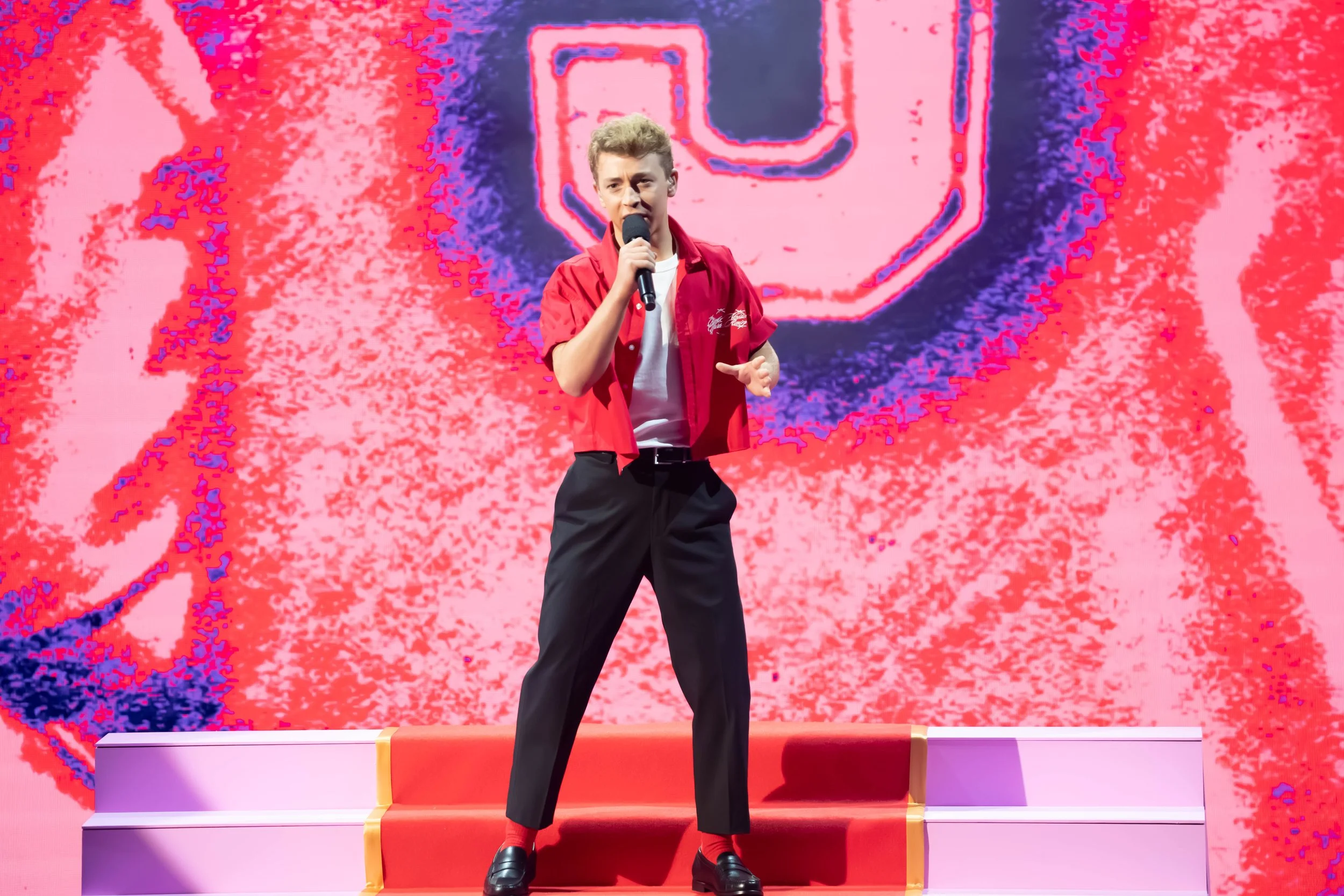 Young man in red short-sleeve shirt, white t-shirt, black pants, and loafers standing on a stage with pink and purple backdrop, holding a microphone.