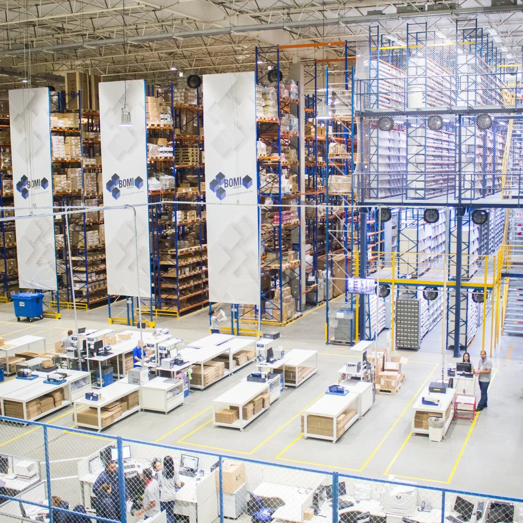 A large warehouse with high shelves filled with boxes and products. There are workers at tables handling packages, and the area is enclosed with safety fencing.