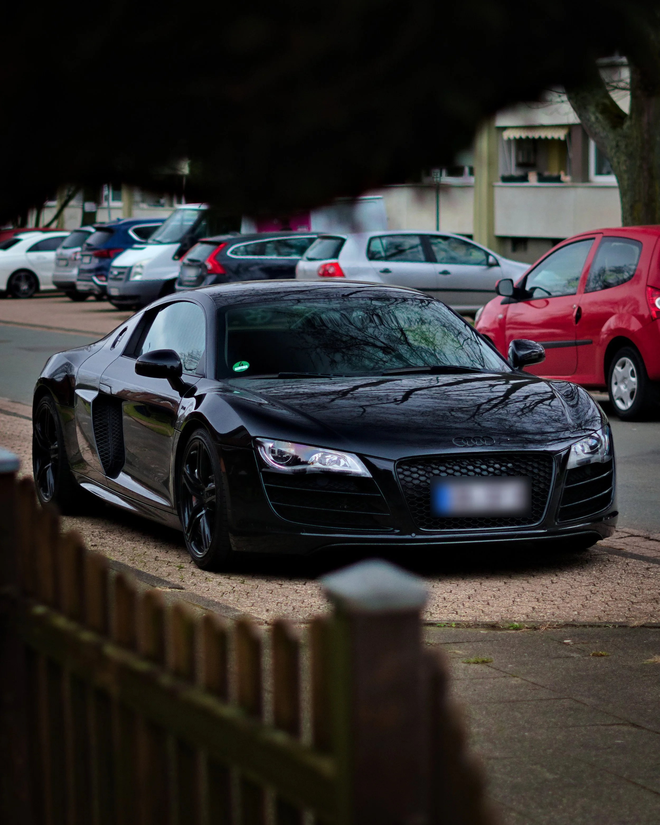 Ein schwarzer Audi R8 Sportwagen auf einem Straßenparkplatz, teilweise durch einen Baum im Vordergrund verdeckt, mit mehreren Autos im Hintergrund.