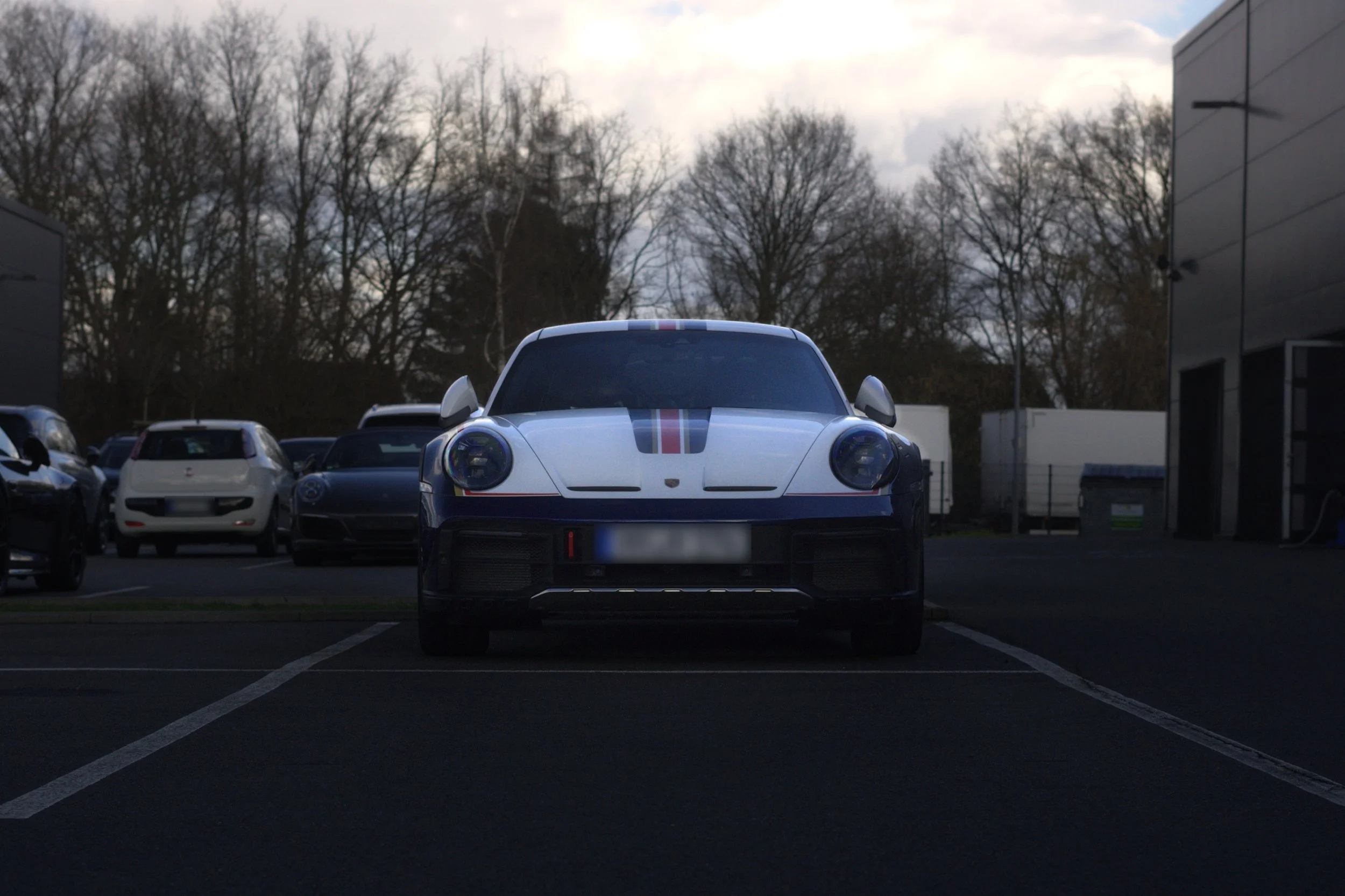 Frontansicht eines weißen Porsche 911 mit schwarzen Streifen auf dem Parkplatz, umgeben von anderen Autos, bei bewölktem Himmel.