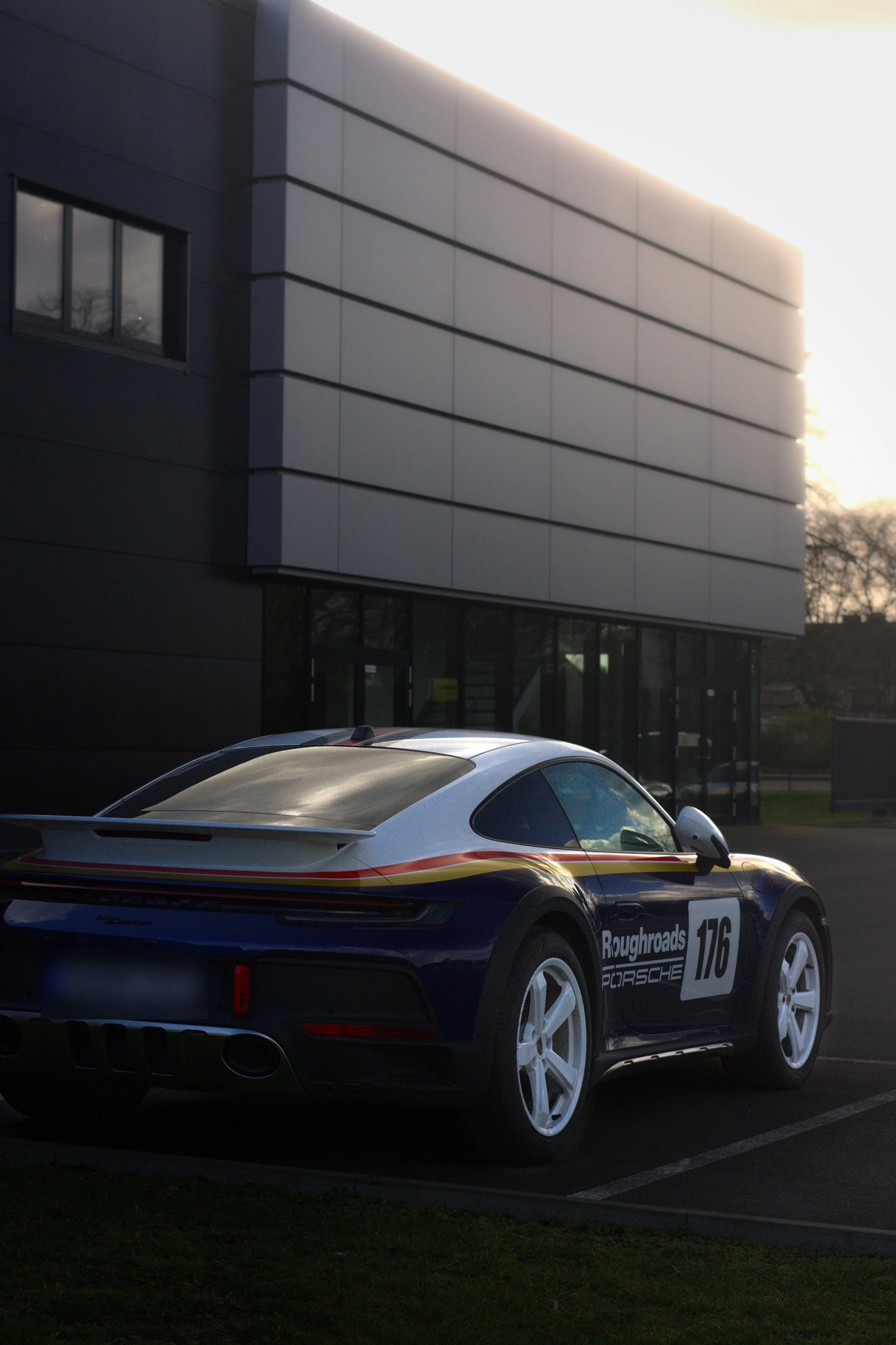 Porsche Rennwagen mit Rennnummer 176 und Sponsorenaufklebern, geparkt außerhalb eines modernen Gebäudes bei Sonnenuntergang.