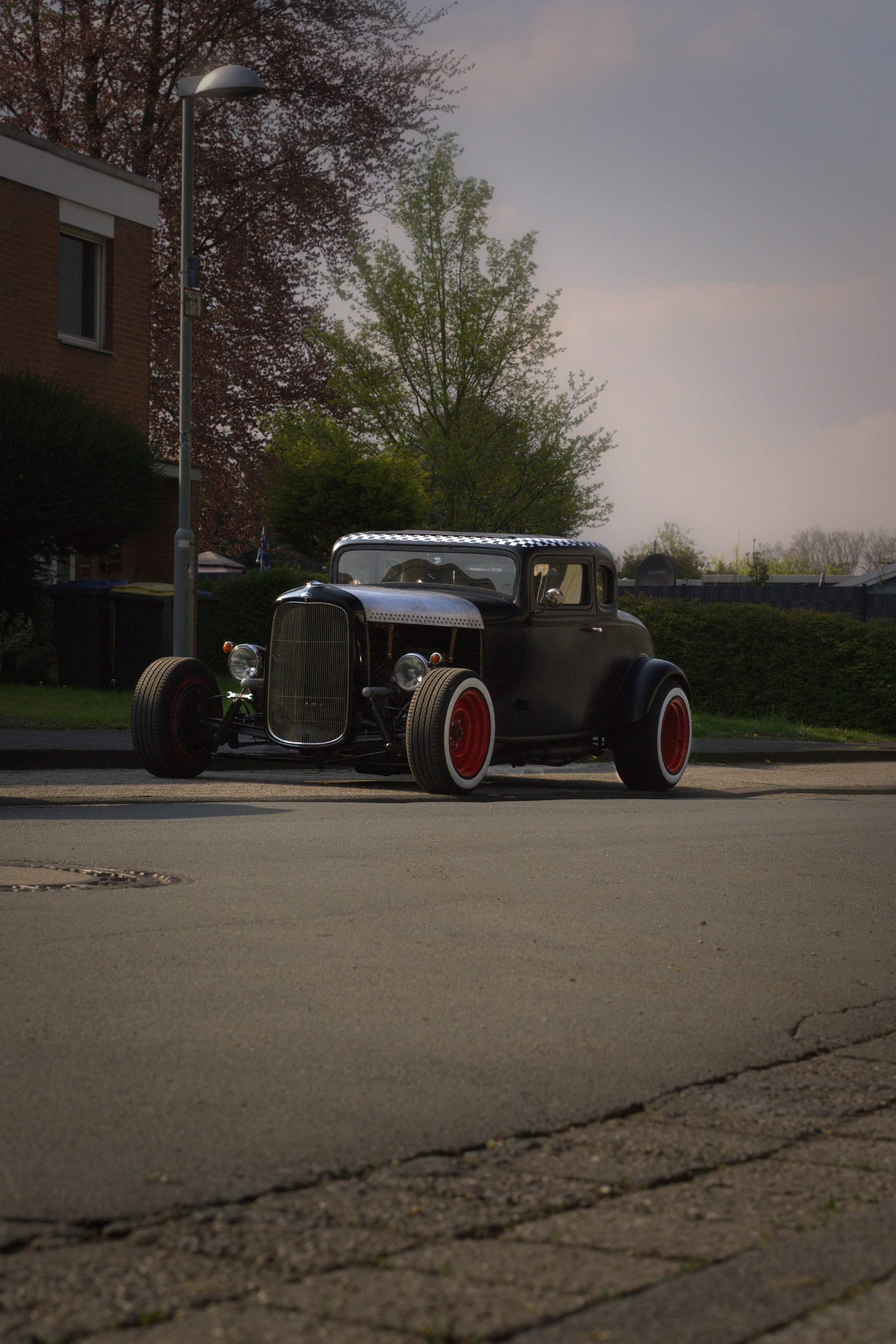 Ein alter, schwarzer Oldtimer mit roten Felgen steht an der Straßenseite, umgeben von Bäumen und Häusern bei Sonnenuntergang.