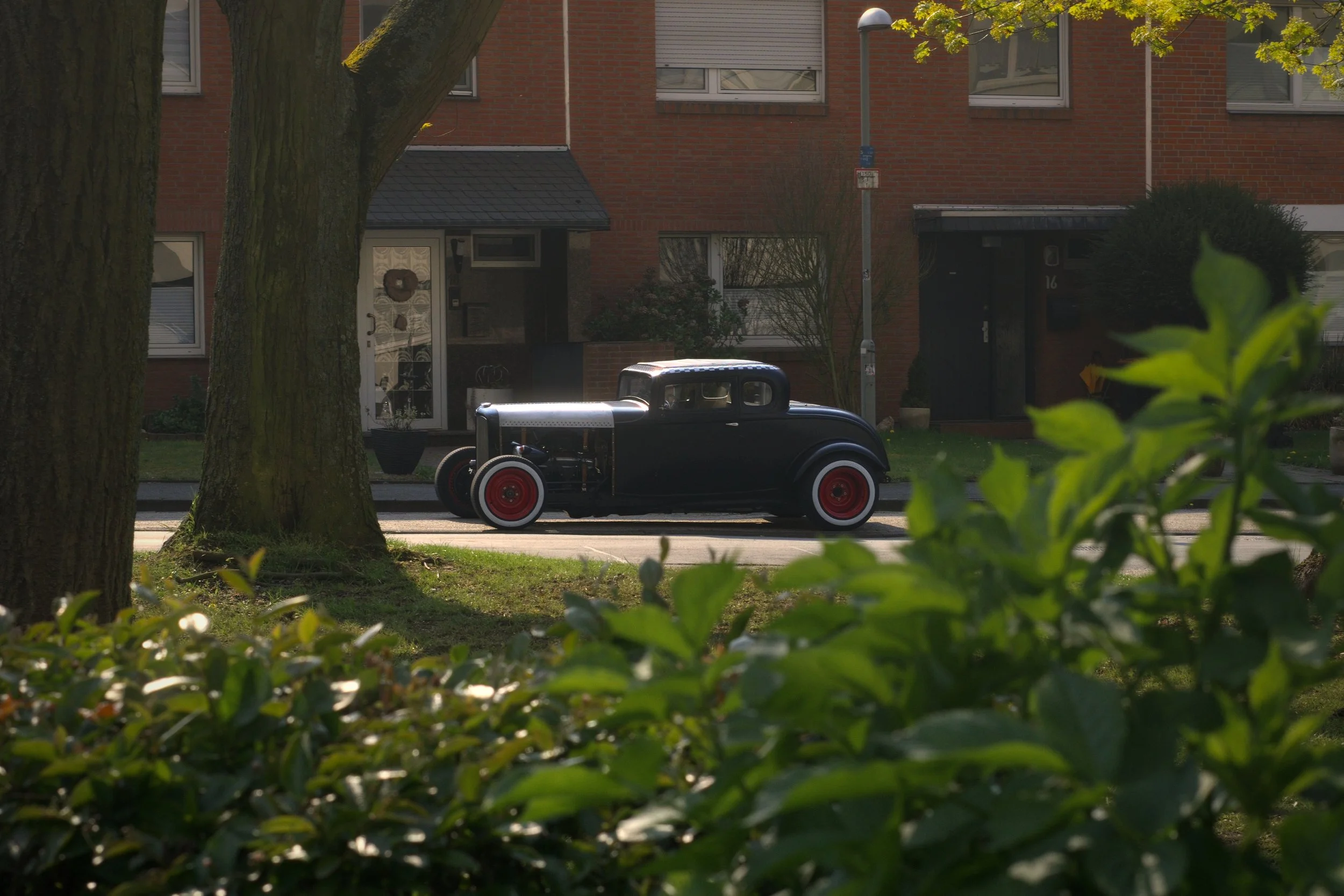 Alter schwarzer Oldtimer mit roten Felgen parkt an einer urbanen Straße, umgeben von Bäumen und Wohnhäusern.