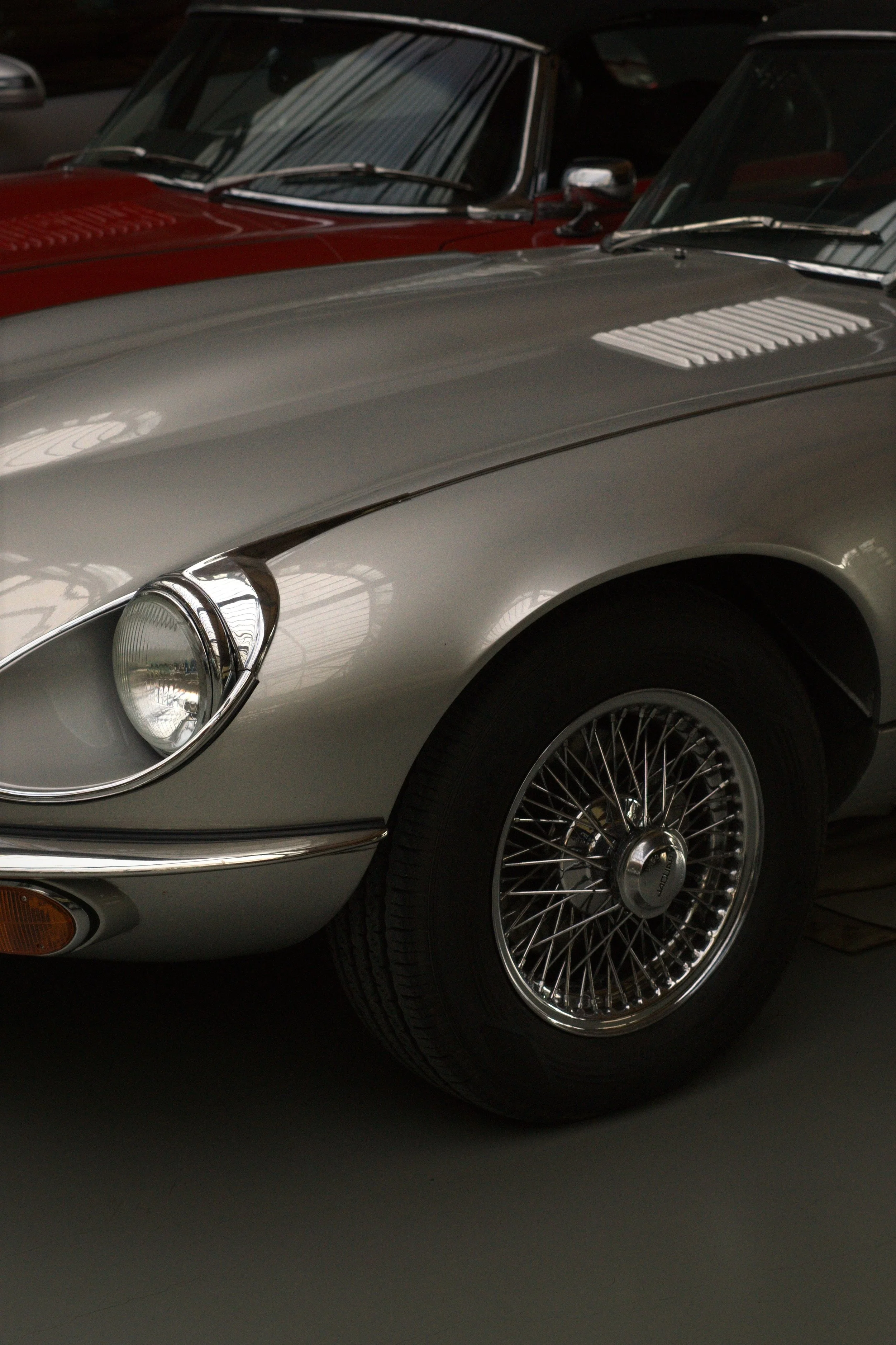 Nahaufnahme eines silbernen Oldtimers, mit sichtbarer radialer Drahtfelge auf dem vorderen Rad. Im Hintergrund ist ein roter Oldtimer sichtbar.