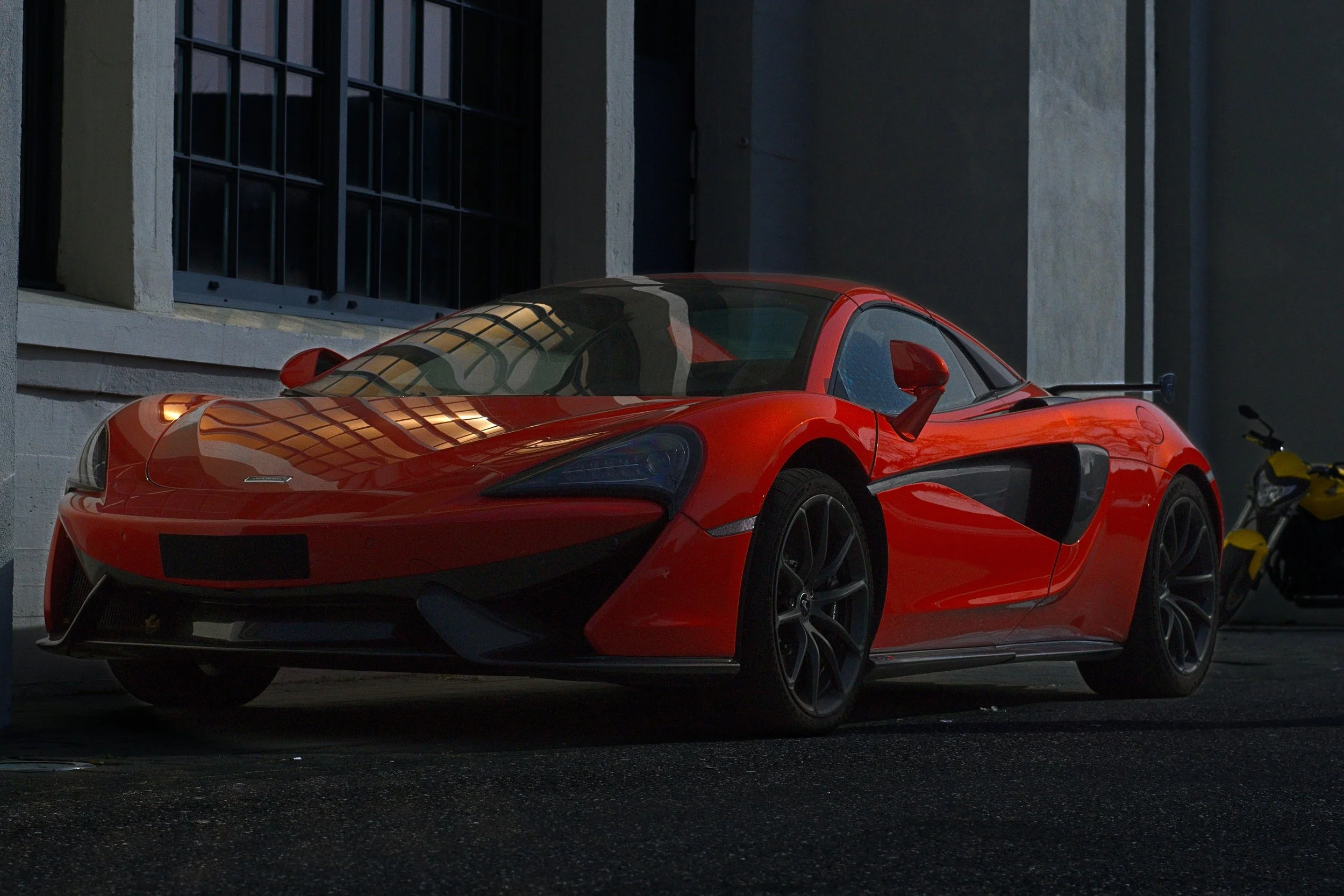 Roter Sportwagen, vermutlich ein McLaren, steht in einer dunklen Garage oder auf einer Straße bei Nacht, reflektierende Lichter spiegeln sich auf der glänzenden Oberfläche.