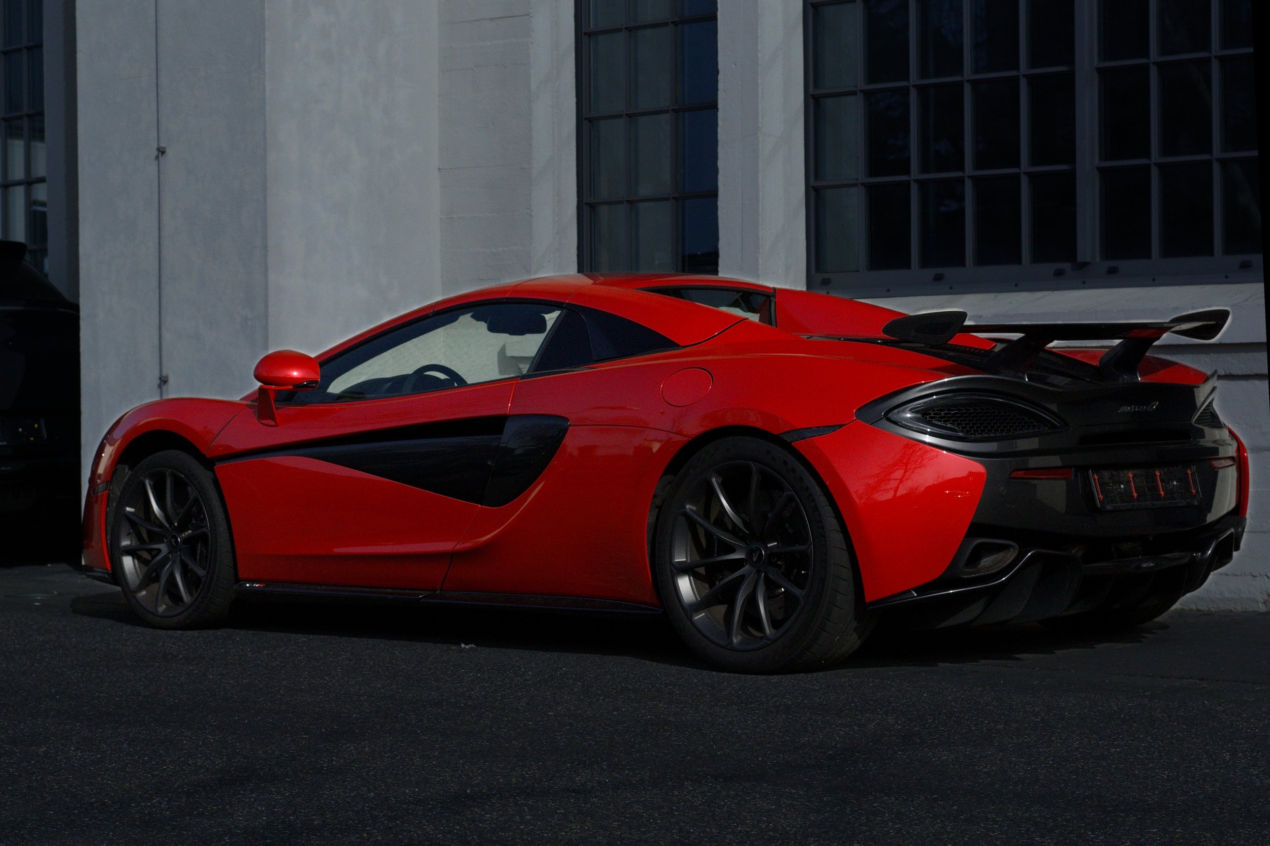 Roter Sportwagen mit schwarzem Akzent, Seitenflügel und Auspuff im Hintergrund eines Gebäudes.