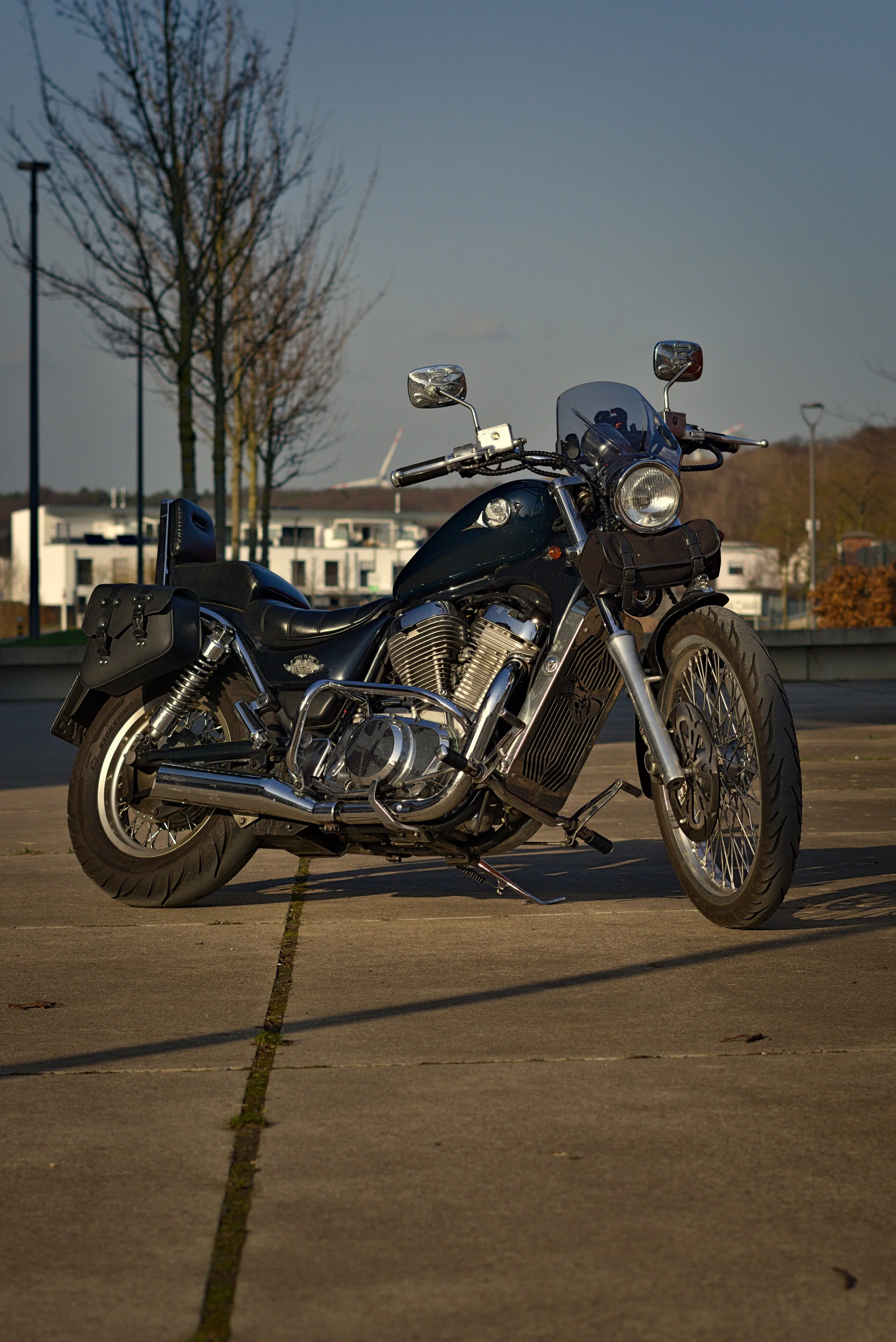 Schwarze Harley-Davidson-Motorrad auf einem Parkplatz bei Sonnenuntergang, mit Stadt und Bäumen im Hintergrund.