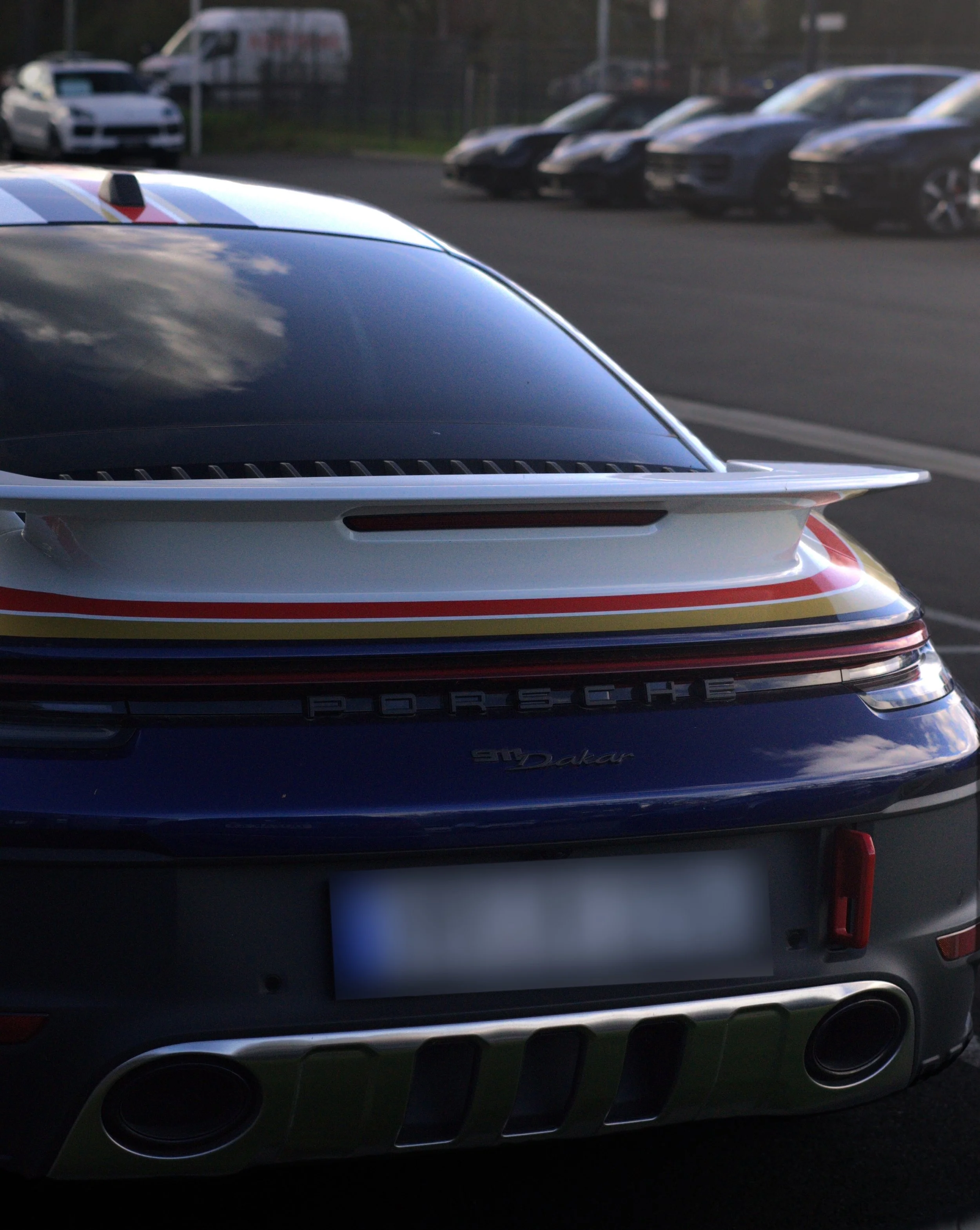 Rückansicht eines Porsche 911 mit Porsche Schriftzug und SD Dakar Branding, Parkplatz mit mehreren Autos im Hintergrund.