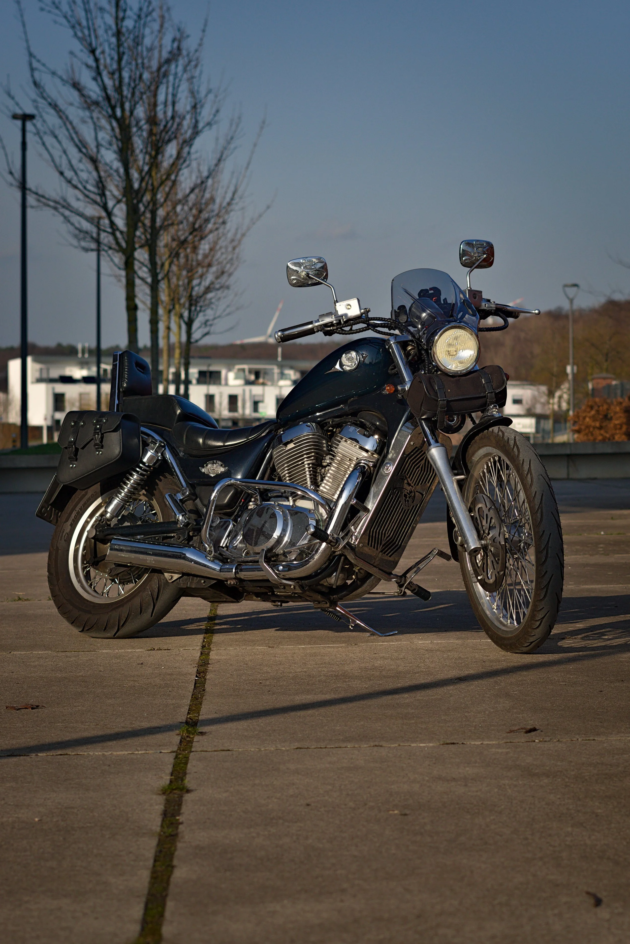 Motorrad auf einem Parkplatz im Freien, mit Bäumen und modernen Gebäuden im Hintergrund, bei Tageslicht.