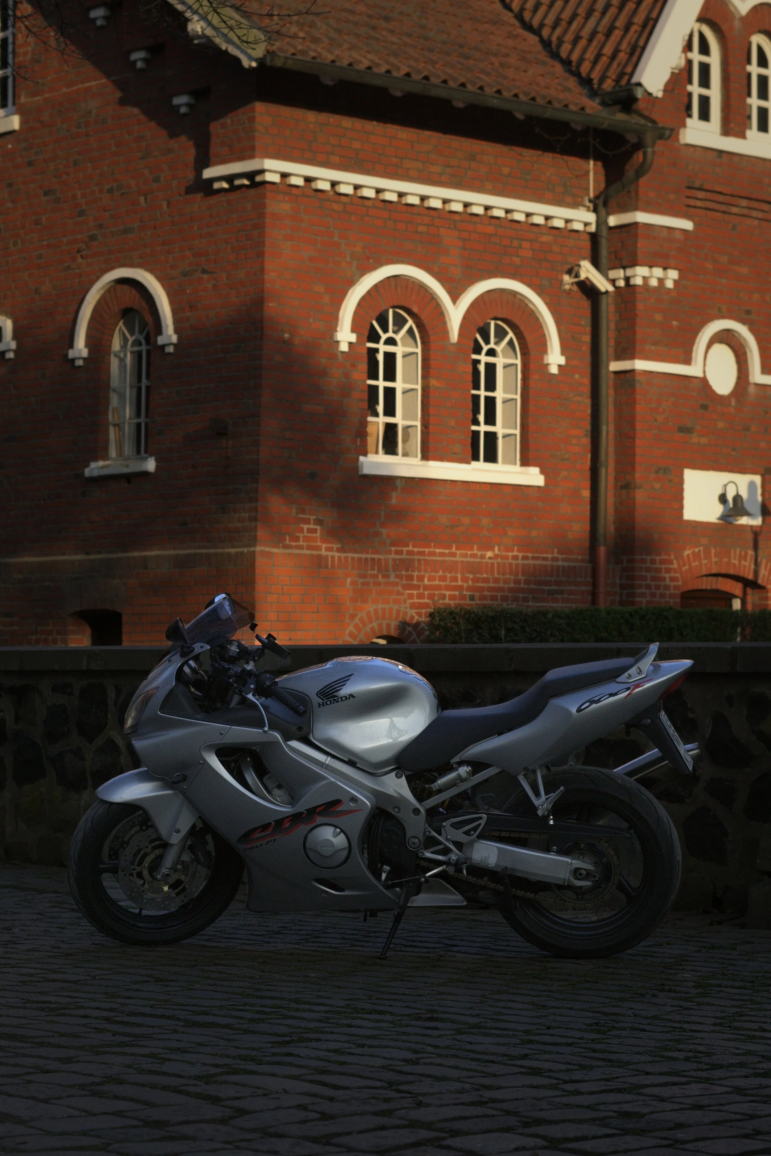 Ein silberner Honda CBR Motorrad steht auf gepflastertem Boden vor einer Backsteinmauer mit historischem Gebäude im Hintergrund, das arched Fenster und weiße Rahmen aufweist.