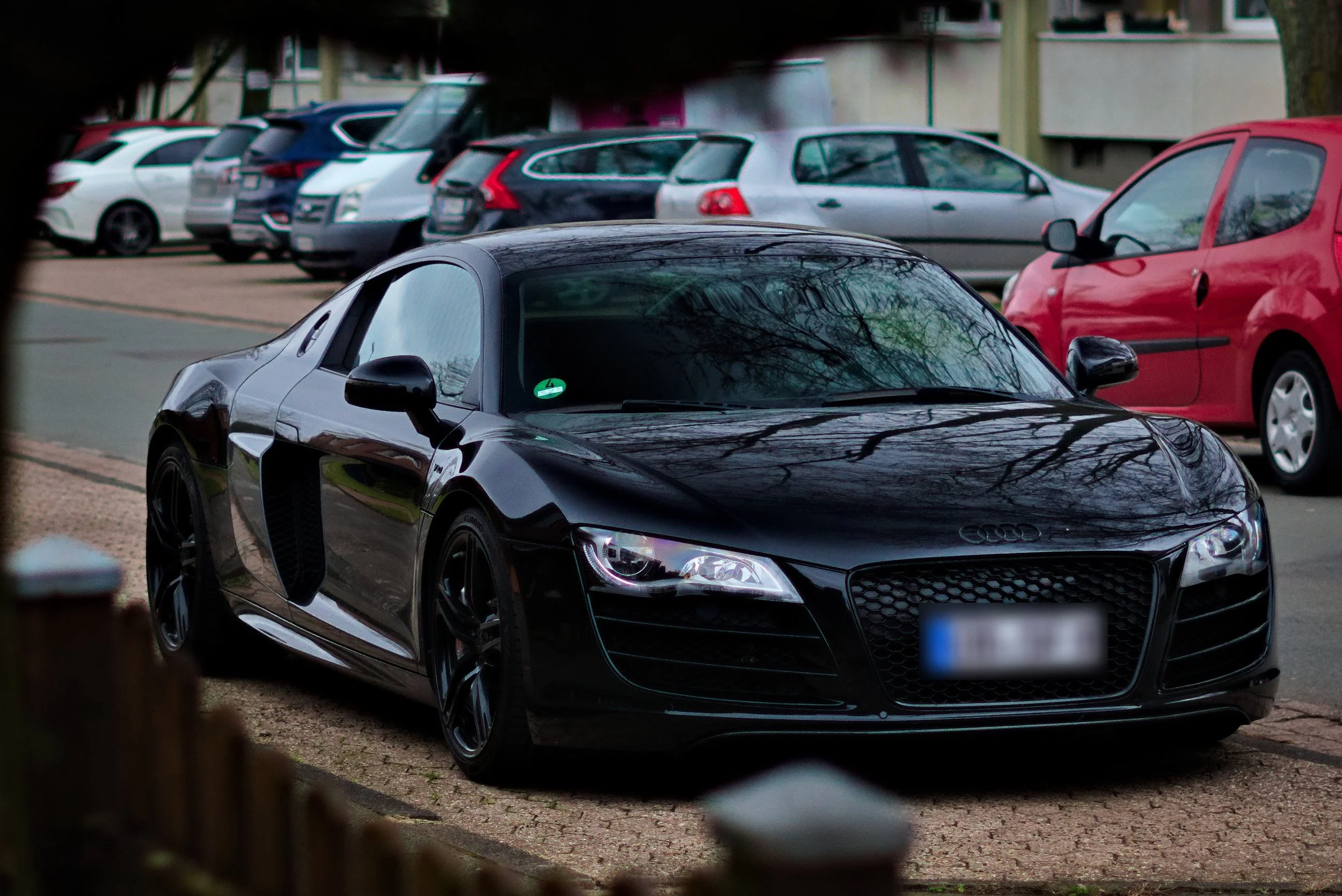 Schwarzer Sportwagen, vermutlich ein Audi R8, geparkt auf einem Parkplatz, umgeben von anderen Autos, mit Bäumen und Gebäuden im Hintergrund.