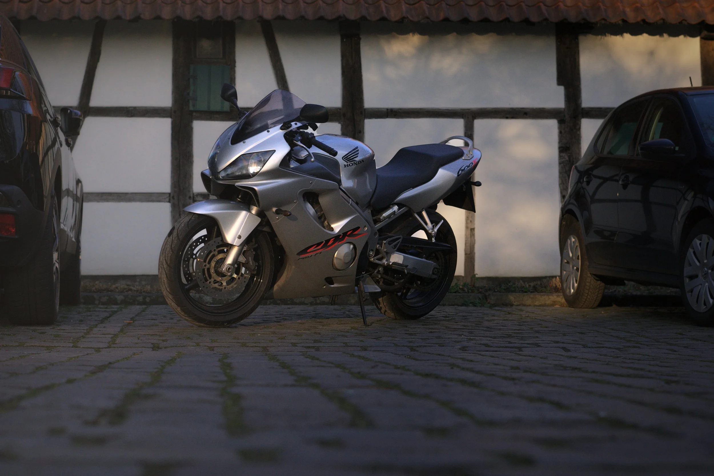 Ein silberner Honda CBR Motorrad steht zwischen zwei schwarzen Autos auf einem gepflasterten Parkplatz vor einem alten Fachwerkhaus.