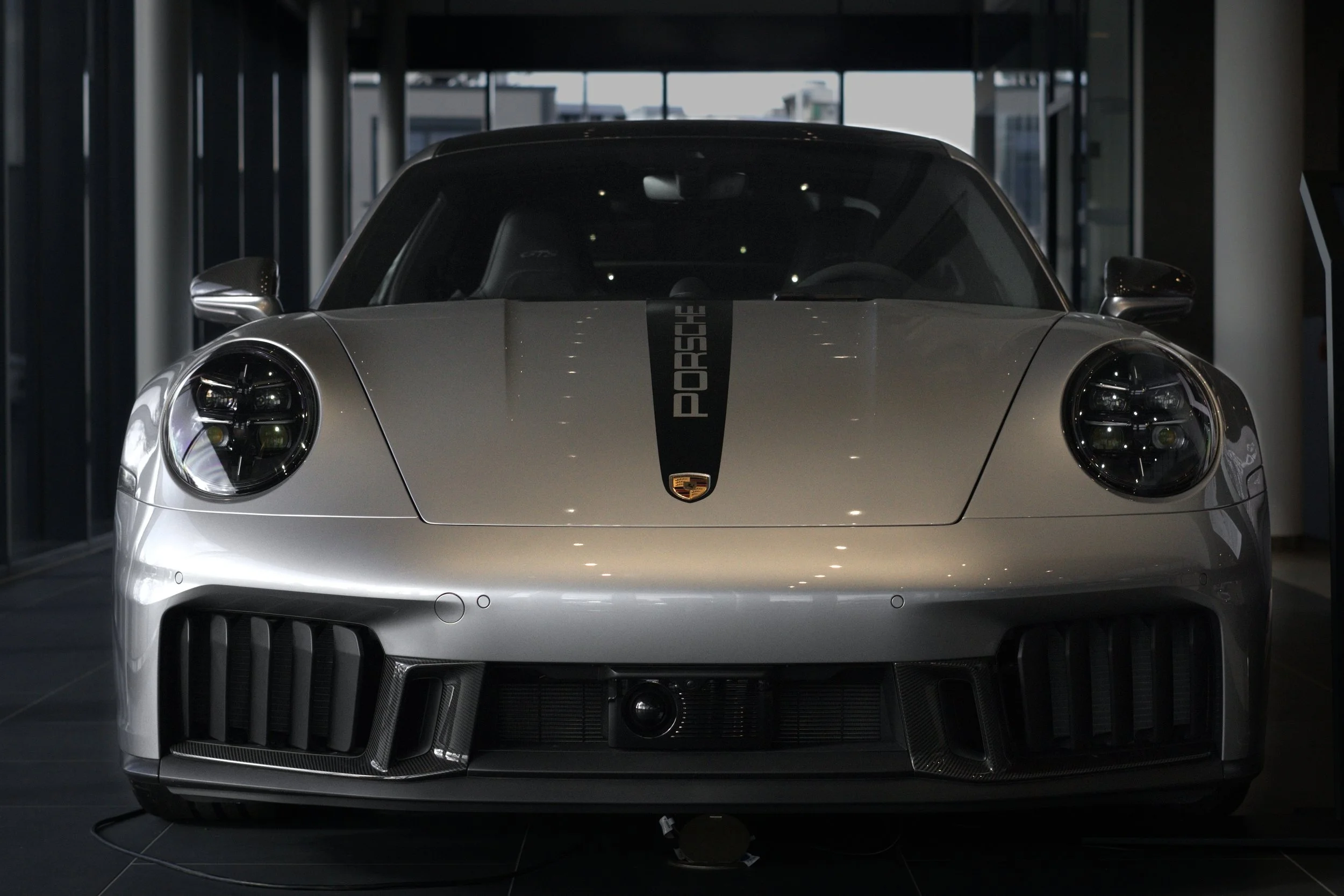Silberner Porsche 911 in einem Autohaus, vorne frontal Ansicht, mit schwarzem Streifen auf der Motorhaube mit Porsche-Logo.