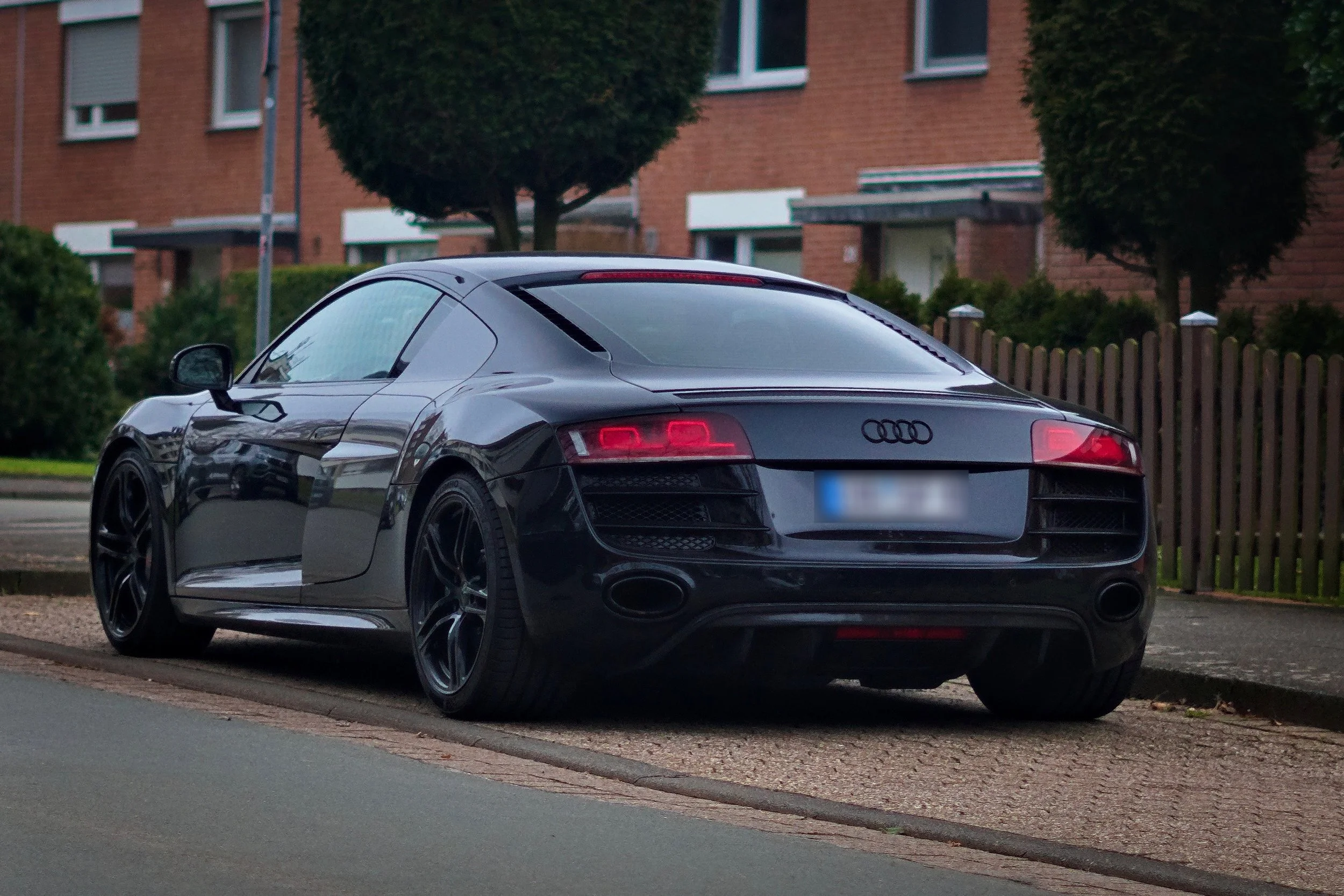 Schwarzer Audi Sportwagen parkend auf dem Gehweg in einer Wohngegend mit roten Backstein-Häusern und Bäumen im Hintergrund.