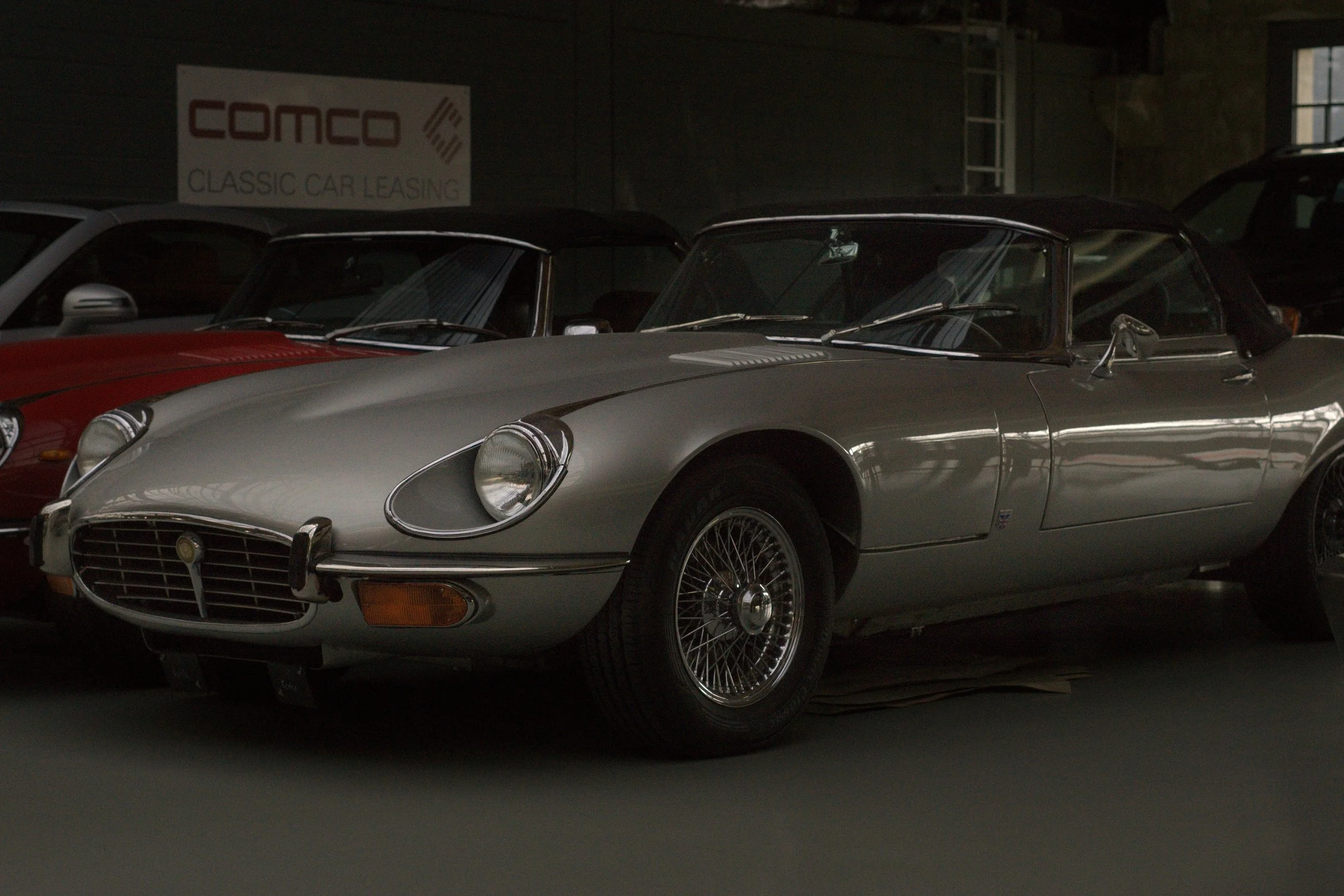 Silberner Oldtimer, vermutlich eine Ferrari 275 GTB, in einem dunklen Raum, vermutlich in einem Autohaus oder Museum, neben einem roten Wagen, im Hintergrund eine Wand mit einem 'comco' Logo.