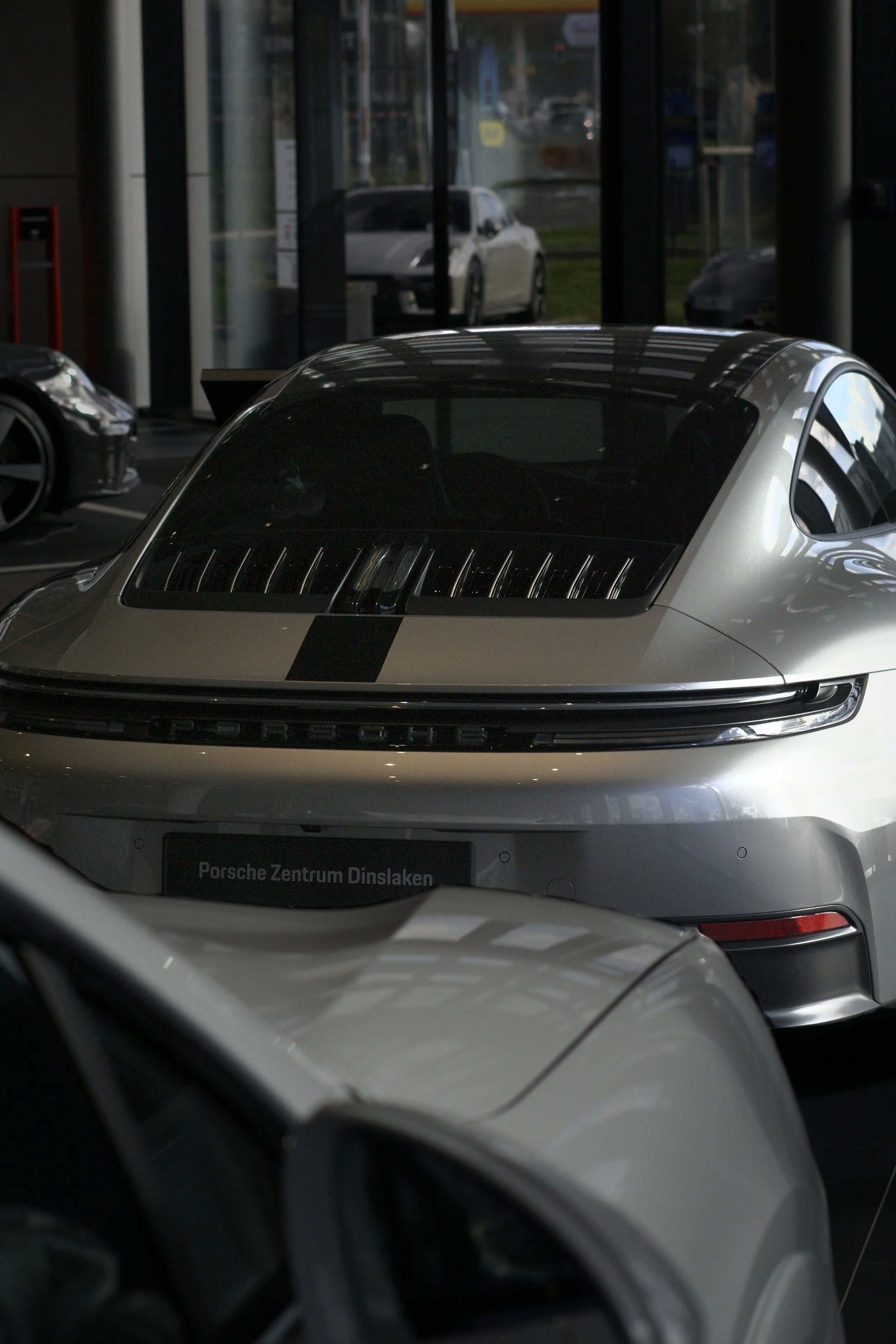 Ein silberner Porsche in einem Autohaus, umgeben von anderen Fahrzeugen, mit Blick durch die Fenster auf einen weißen Pkw im Außenbereich.