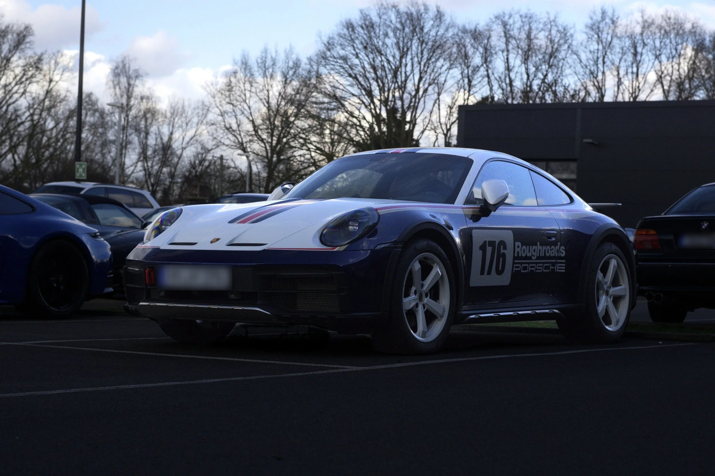 Ein Porsche 911 mit Rennkennzeichen und Aufklebern steht auf einem Parkplatz, umgeben von anderen Autos, bei bewölktem Himmel und kahlblättrigen Bäumen im Hintergrund.