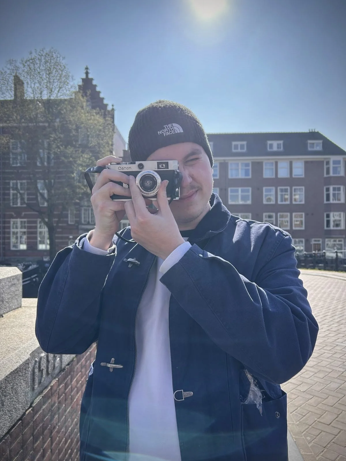 Junger Mann mit schwarzen The North Face Mütze und dunklem Jacke hält eine Kamera vor das Gesicht, während er eine Fotosession im Freien an einem sonnigen Tag macht.