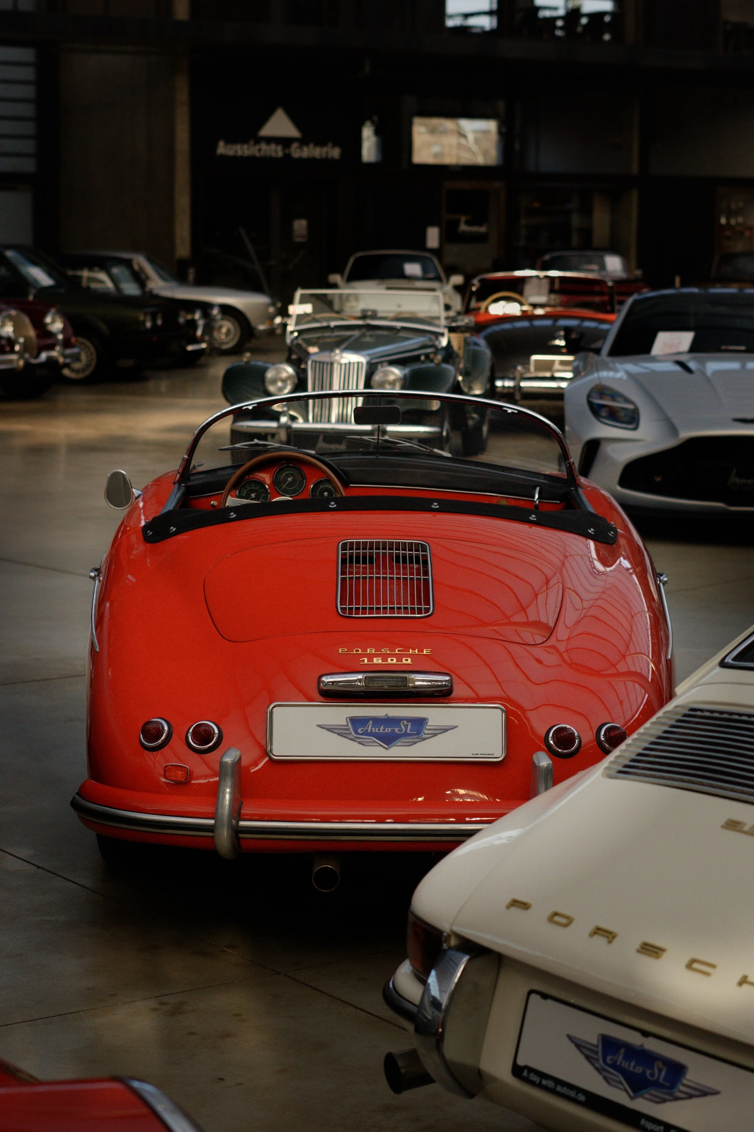 Roter Porsche 356 mit offenem Verdeck in einer Oldtimersammlung, im Hintergrund weitere klassische Autos.
