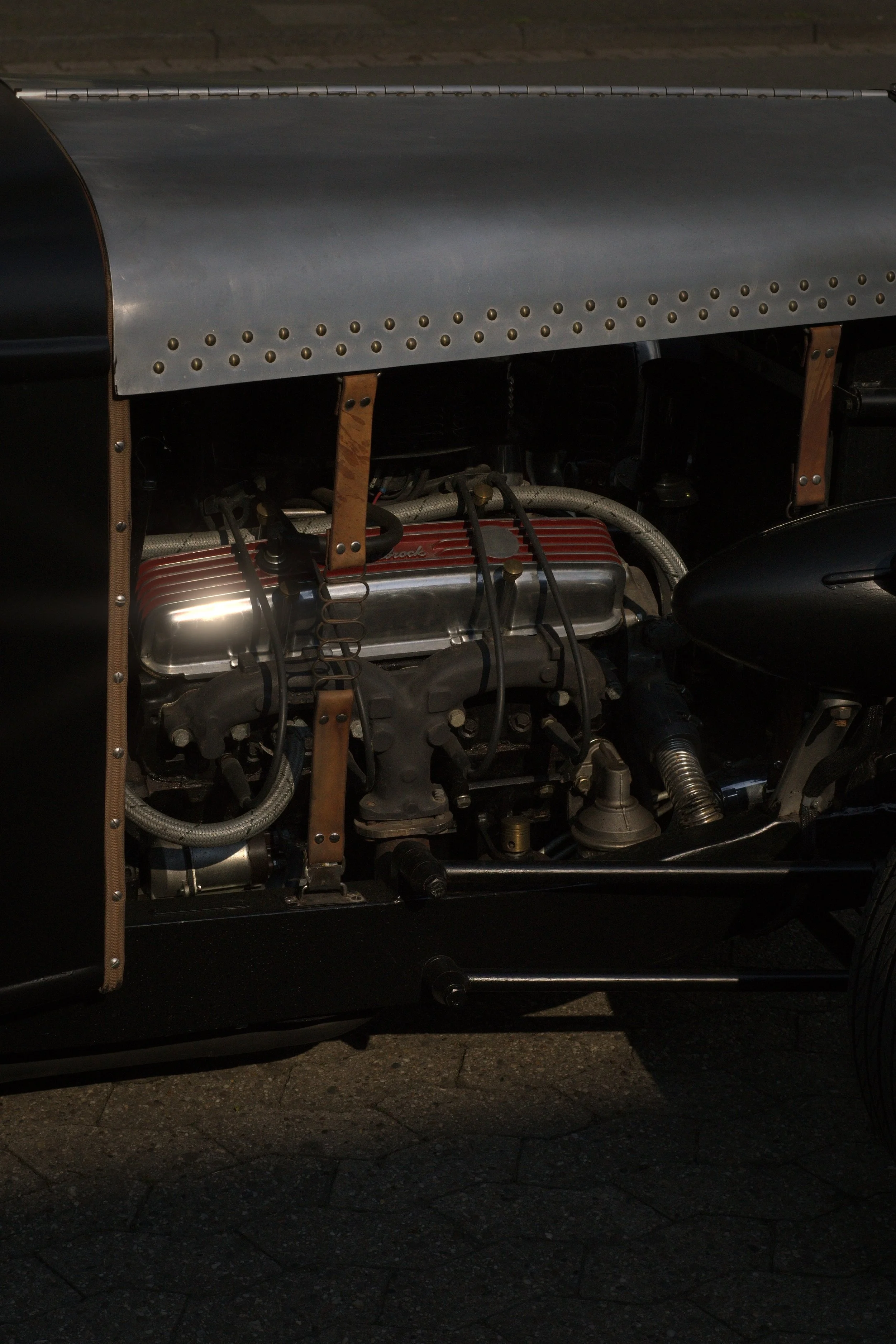 Nahaufnahme eines Automotormotors in einem klassischen Fahrzeug, sichtbar unter einem Metallblech, mit sichtbaren Schrauben, Schläuchen und mechanischen Teilen.