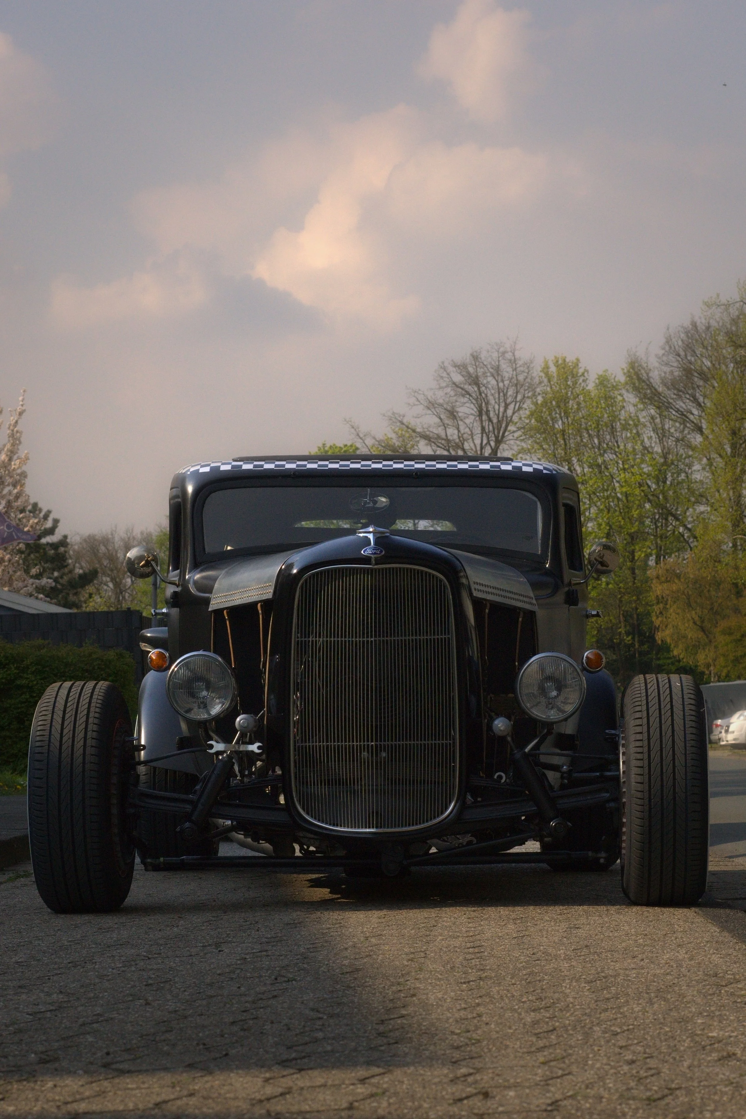 Ein schwarzer, vintage Automobil vor einem Baumpark, Blick von vorne, mit großem Grill und runden Scheinwerfern.