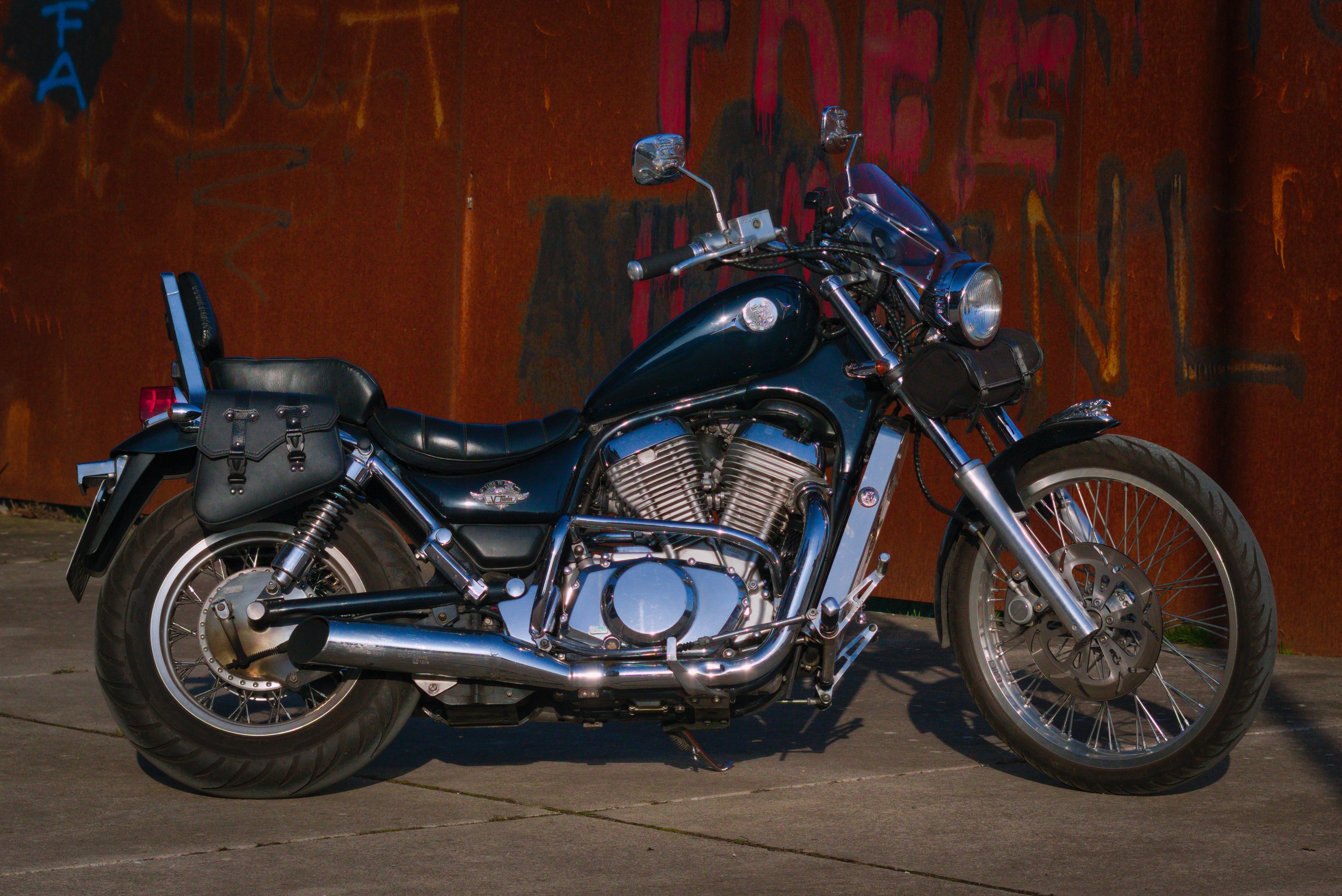 Schwarz lackierte Harley Davidson Motorrad mit Lederkoffern, vor einer rostigen Metallwand mit Graffiti