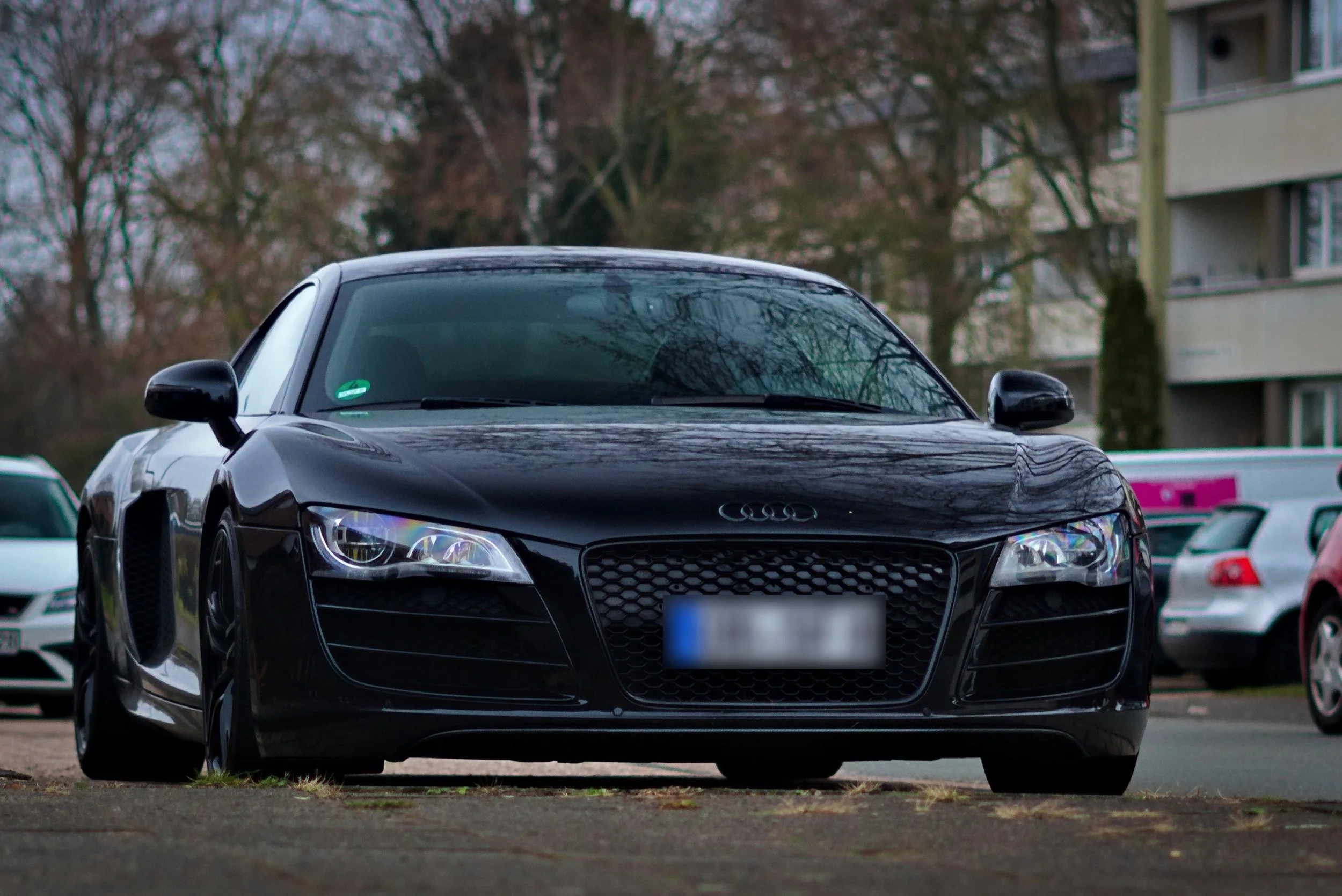 Schwarzer Audi Sportwagen auf einem Parkplatz, umgeben von weiteren Autos, im Hintergrund Bäume und ein Mehrfamilienhaus.