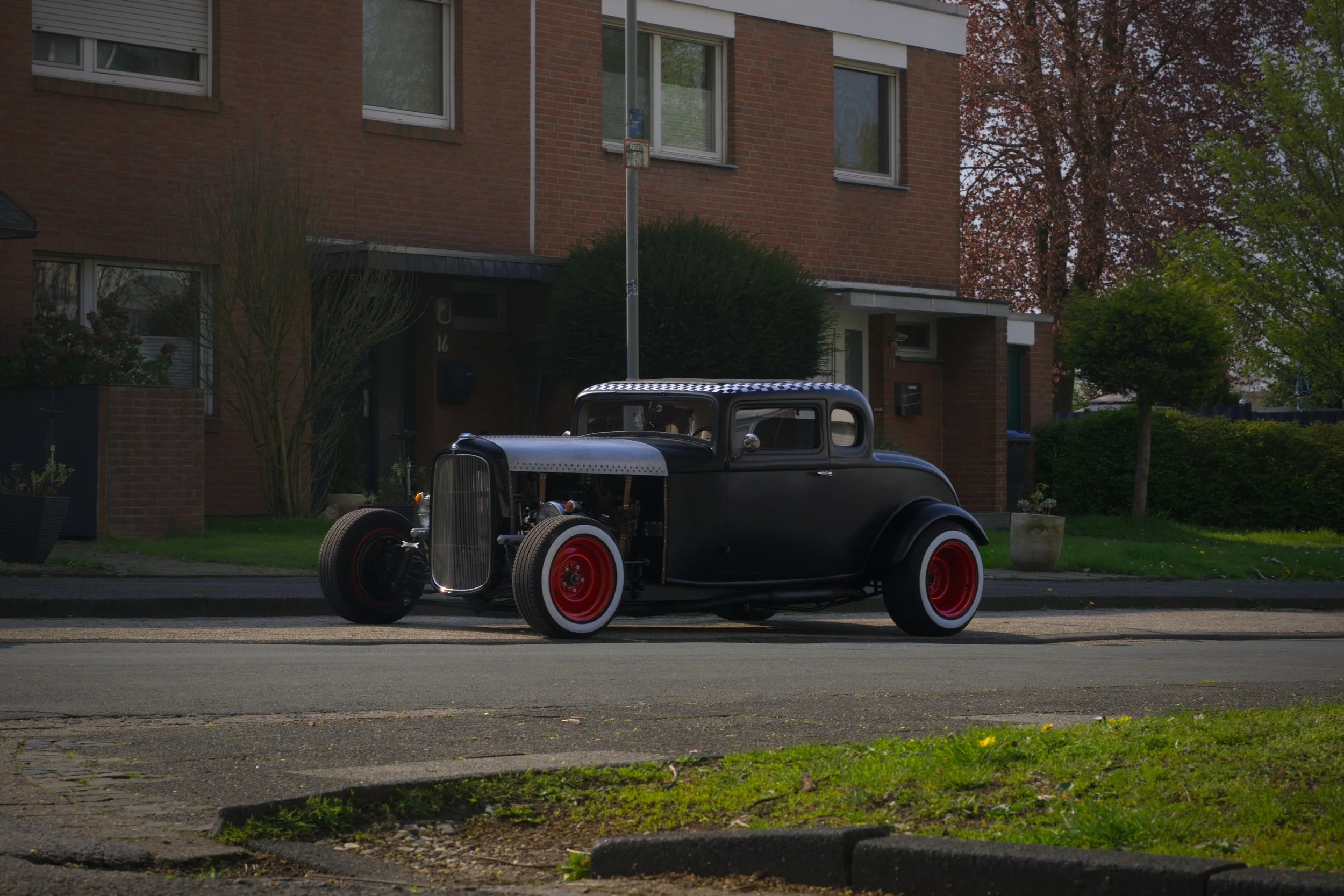Ein schwarzes, klassisches Auto mit roten Felgen steht auf der Straße vor einem modernen Wohnhaus. Das Auto ist teilweise offen, ohne Kühlergrill und Motorhaube.