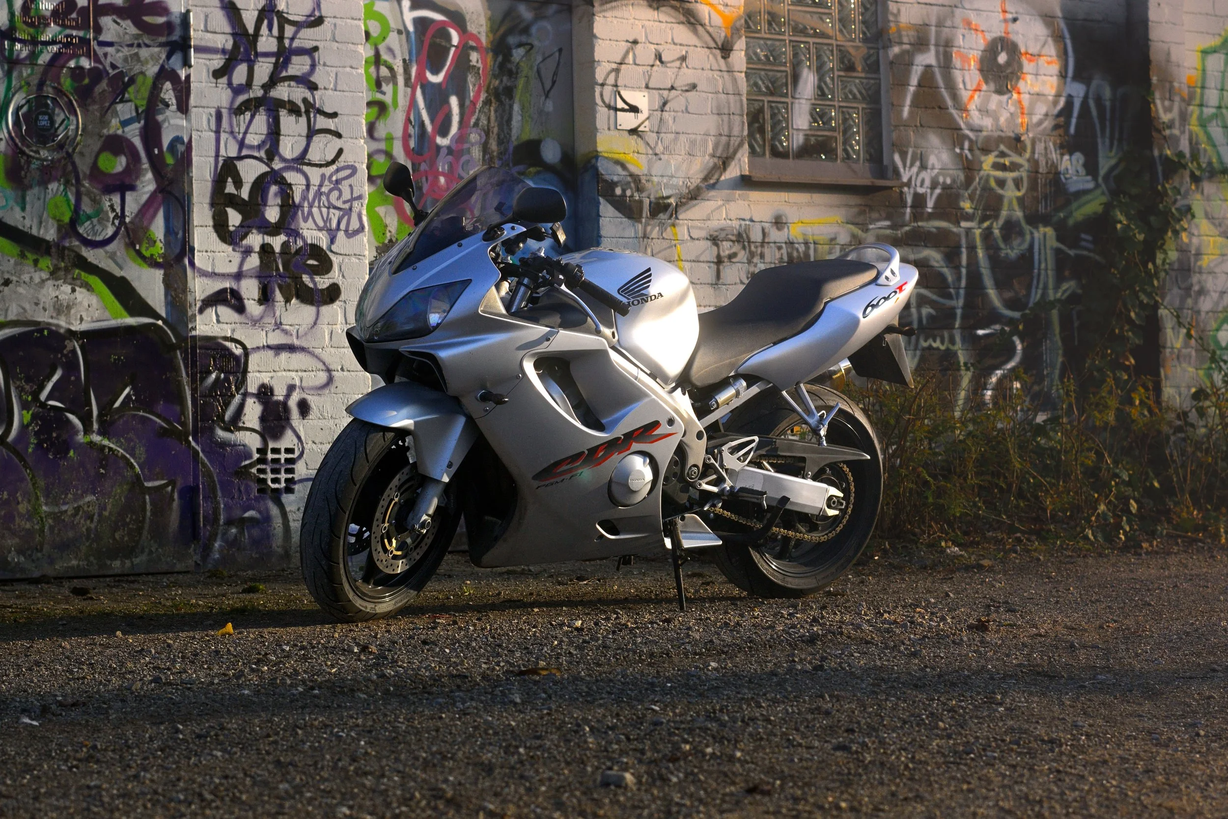 Ein silberner Honda CBR Motorrad steht vor einer Wand mit bunten Graffiti. Der Hintergrund zeigt eine verputzte Wand mit Graffiti und einem kleinen Fenster.